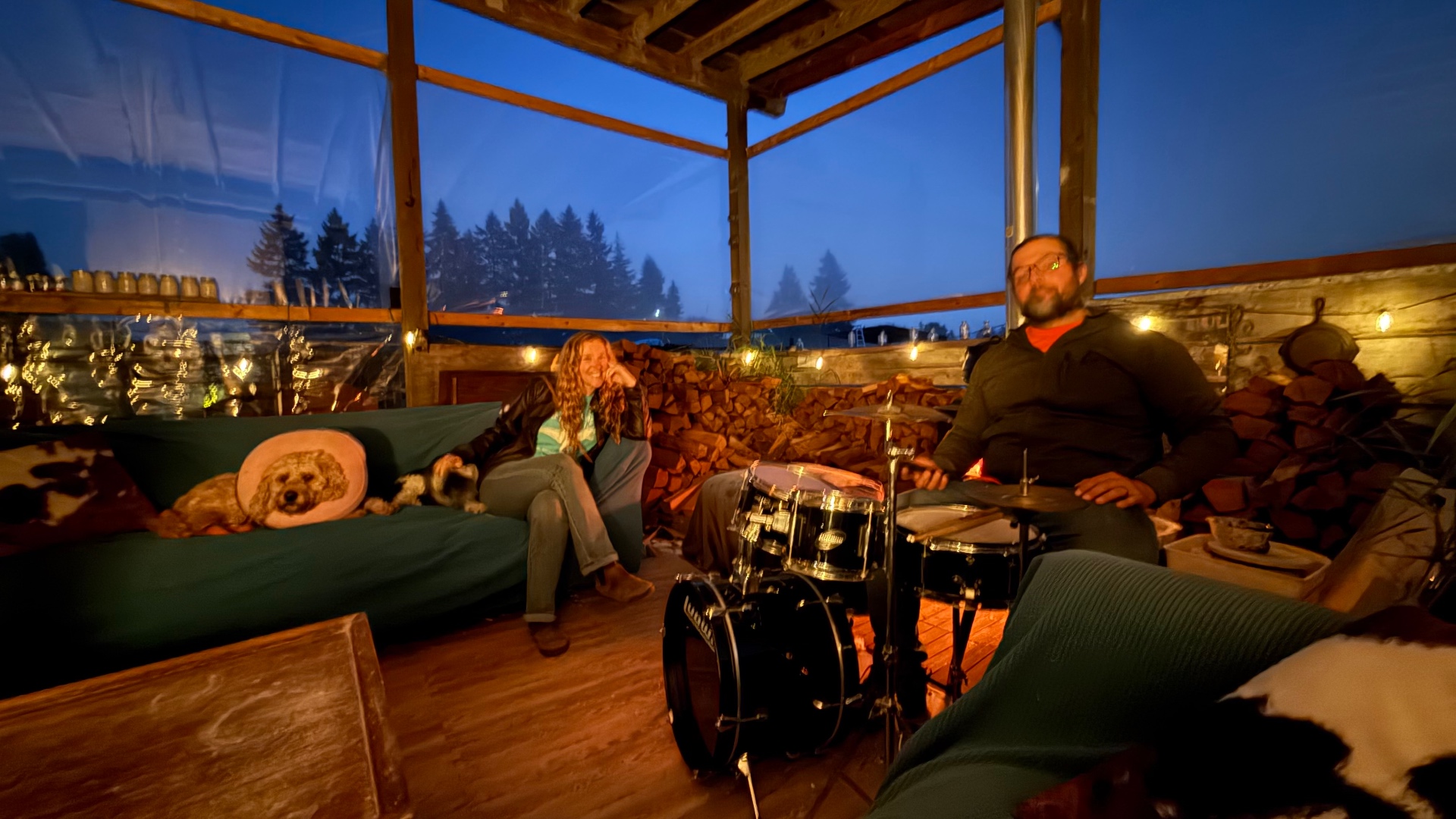 Two friends hanging out together on an enclosed porch at twilight — a woman on a teal sofa and a man at a Ludwig drum kit — warmed by string lights and the glow of a fire, a small dog resting nearby