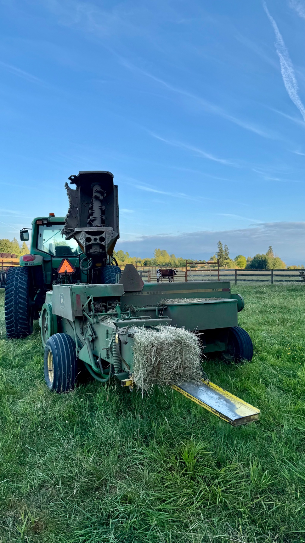 A green John Deere tractor hitched to a hay baler dropping a rectangular bale in a green field under a blue sky, with a cow visible behind a fence