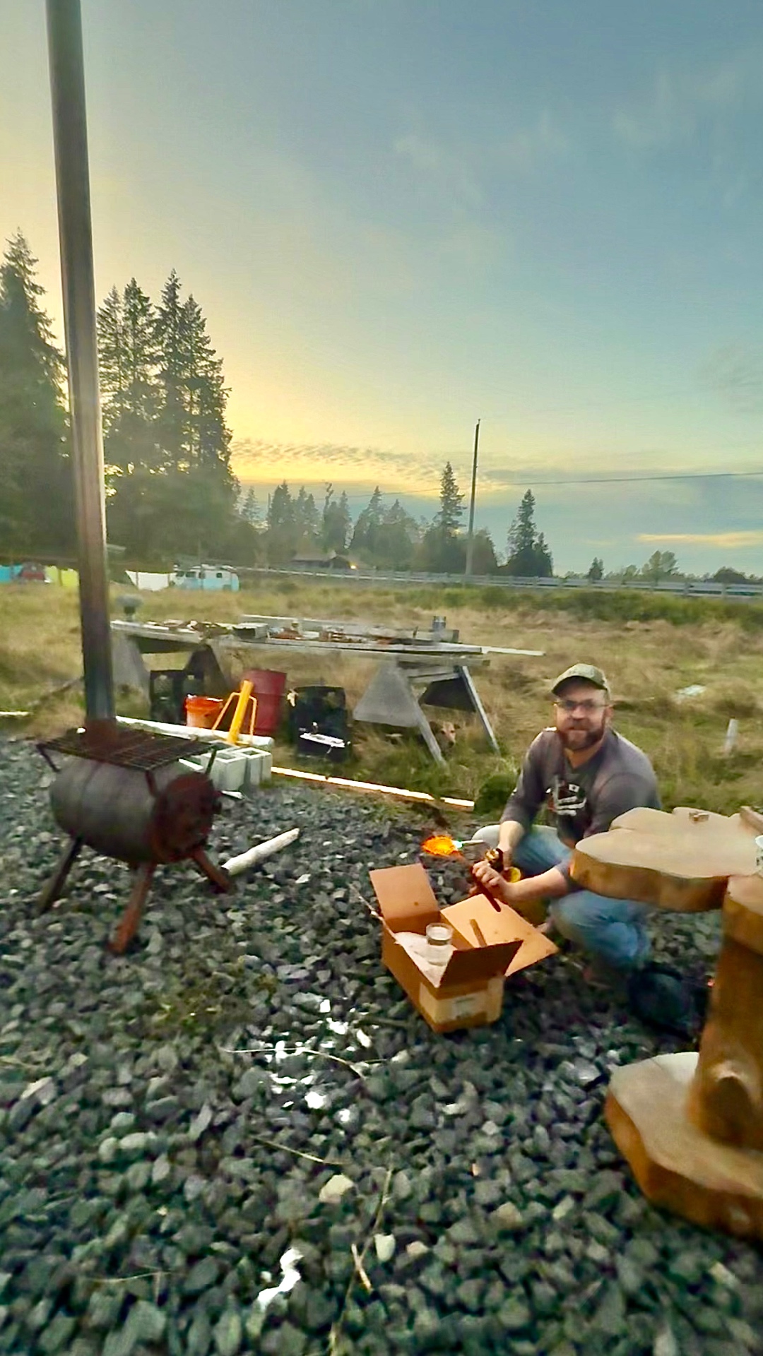 Zach kneeling on gravel at sunset with a torch in hand, a glowing hot piece of metal in his tongs, an outdoor stove and work tables behind him