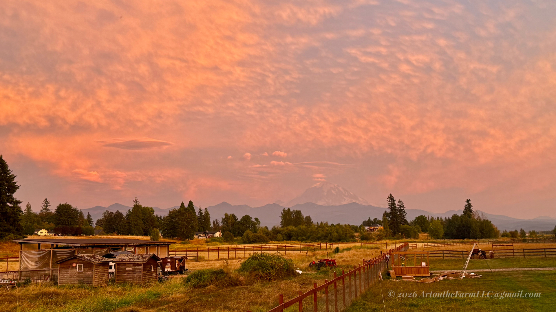 A wide farm scene under a deep orange-pink sunset sky, with weathered outbuildings, a red tractor, a wooden fence, golden fields, and a distant snow-capped peak