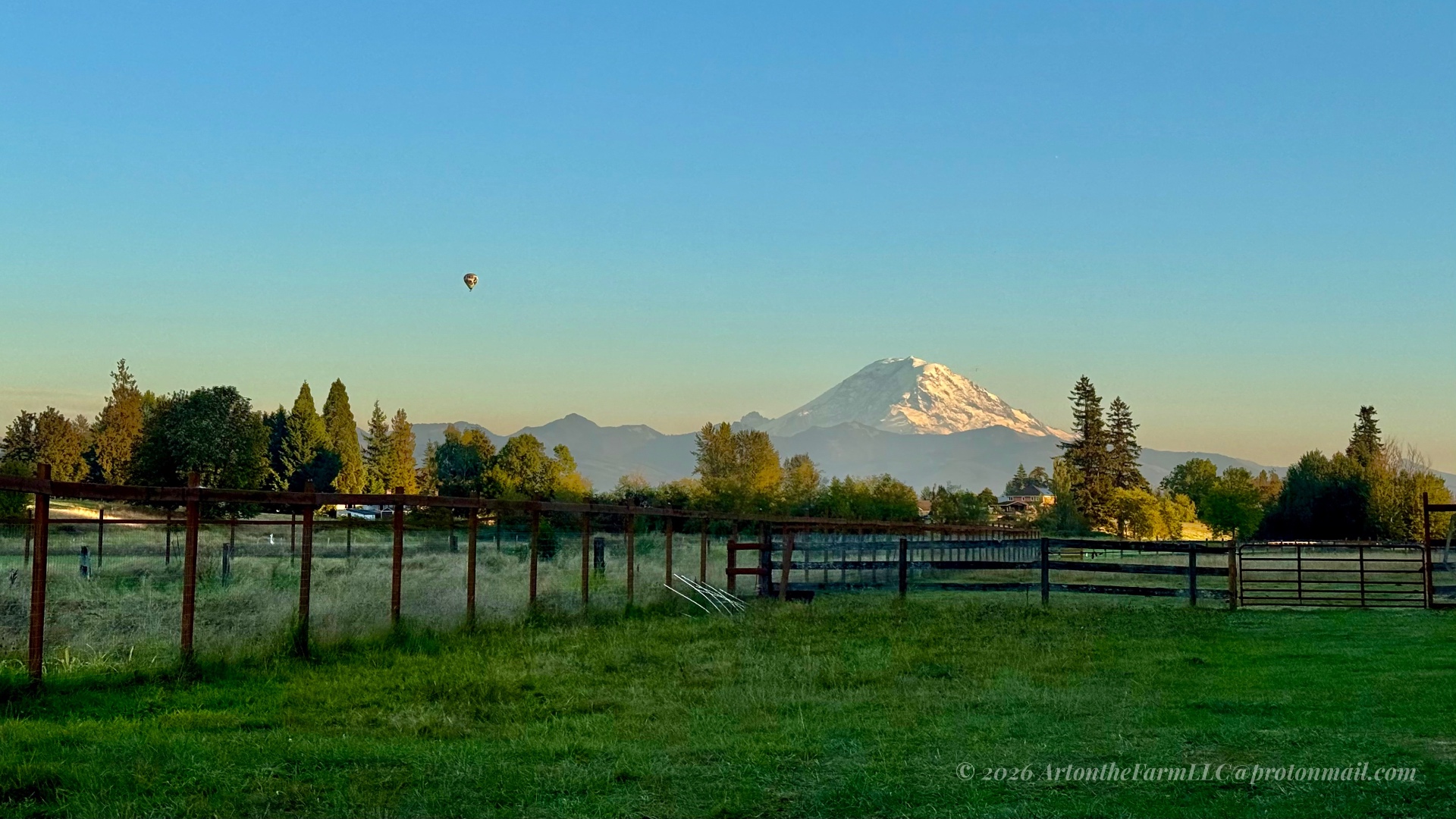 A wide farm field divided by a wooden and wire fence, snow-capped Mount Rainier in the background, and a single small hot air balloon in a clear blue sky
