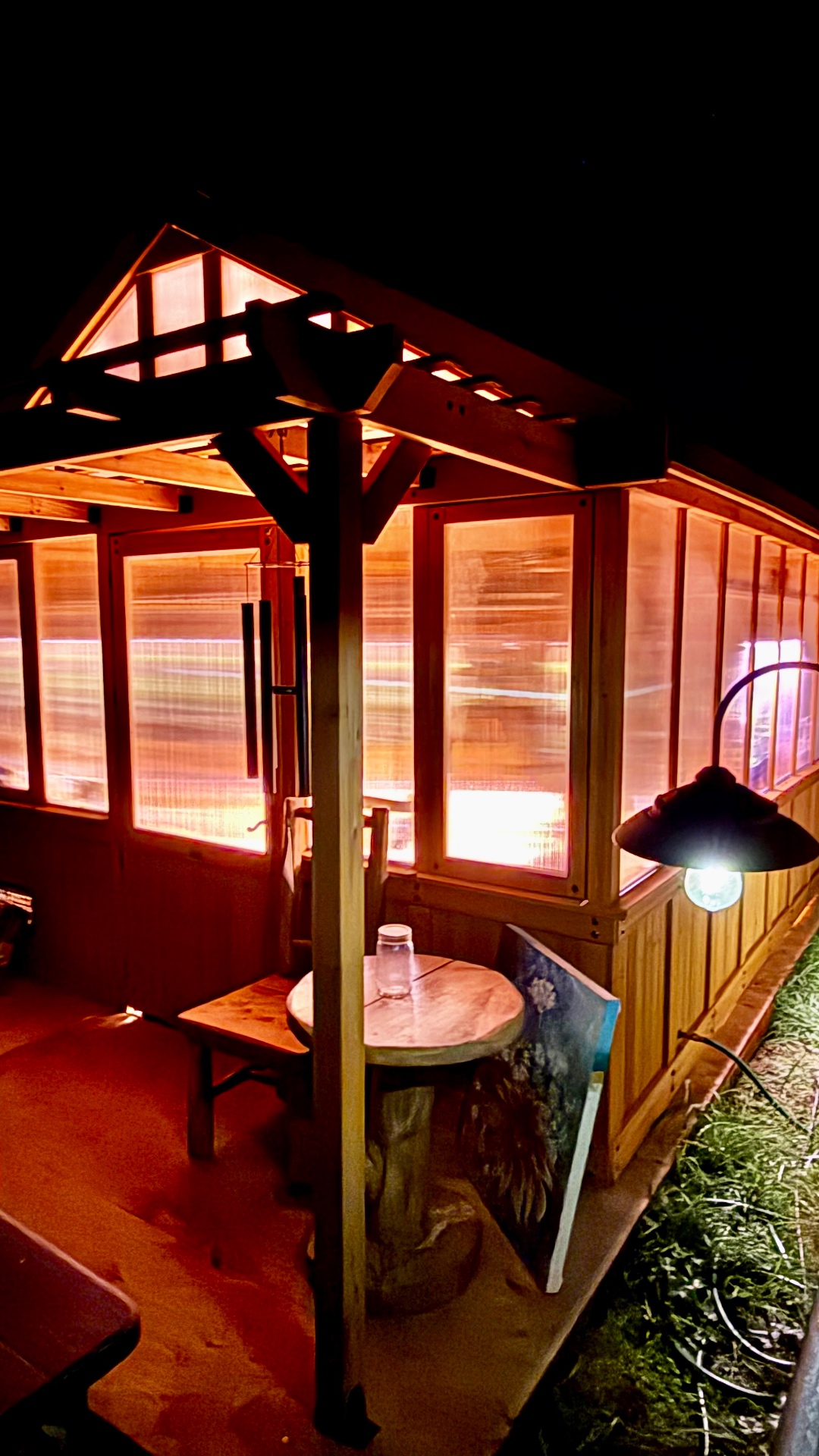 The wooden porch beside the greenhouse at night, with a small round table and a glass jar, a wooden chair, a painted flower canvas leaning against the table, wind chimes overhead, and a warm bulb shining down on the grass