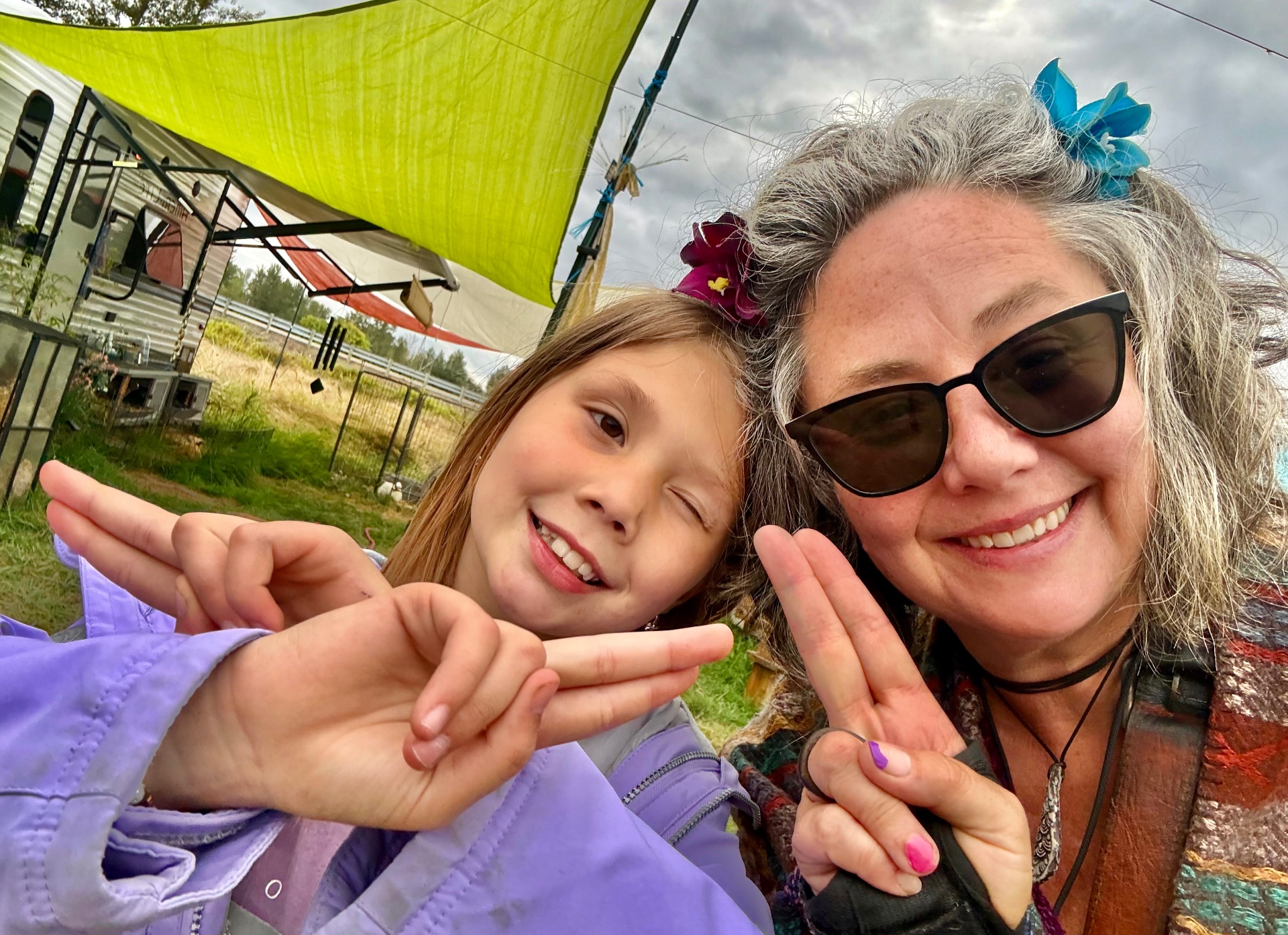 Miss Iz and her young student smiling side by side outdoors on the farm, both flashing peace signs under a bright green shade sail
