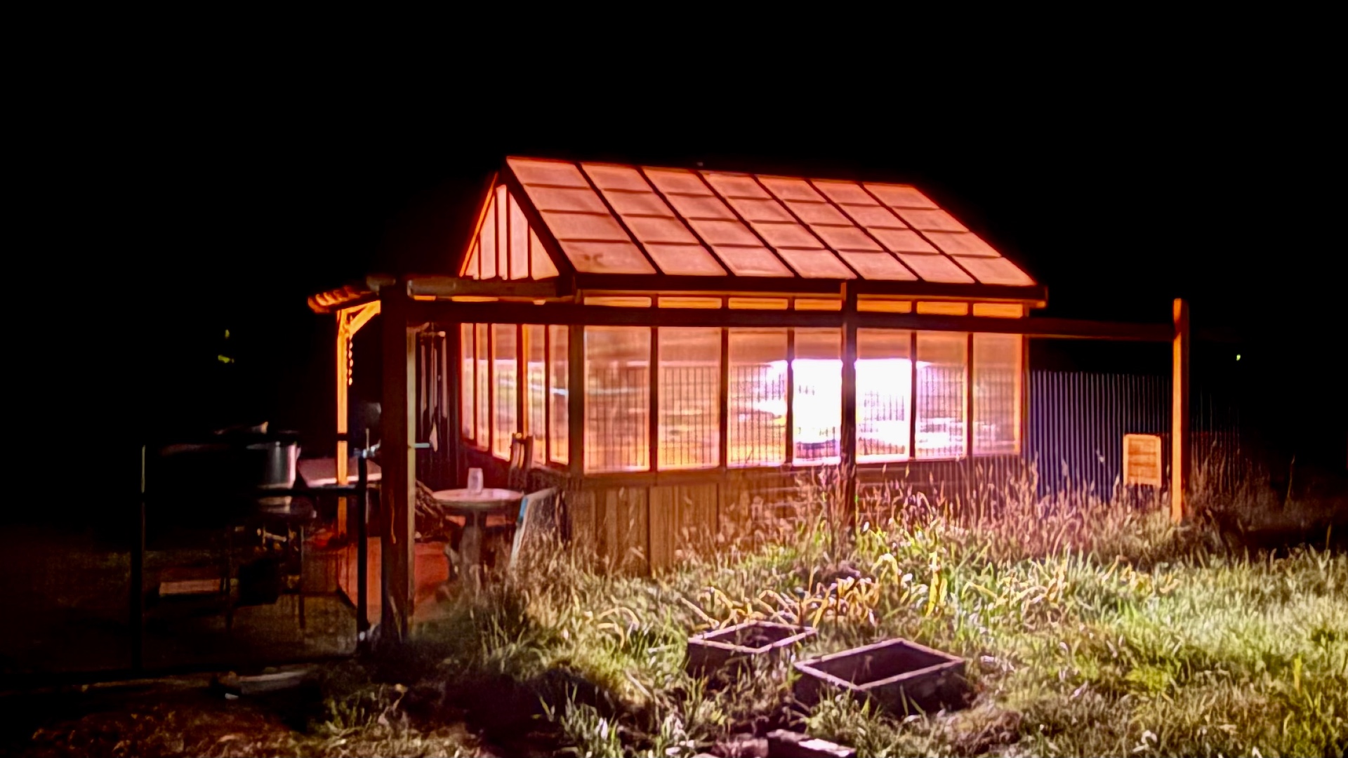 A wooden greenhouse at night, its roof and translucent wall panels glowing warm amber from the lights inside, with cool white grow lights shining through the lower windows and tall grass lit in front