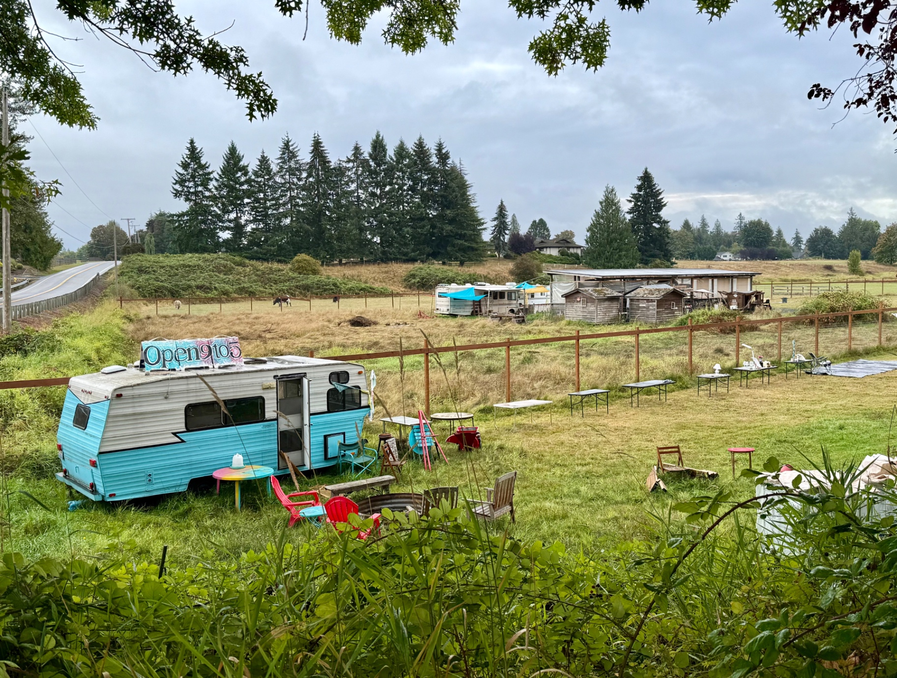 An elevated view of a grassy field with a turquoise trailer and rows of white folding tables, chairs, a fire pit and goods set out for sale