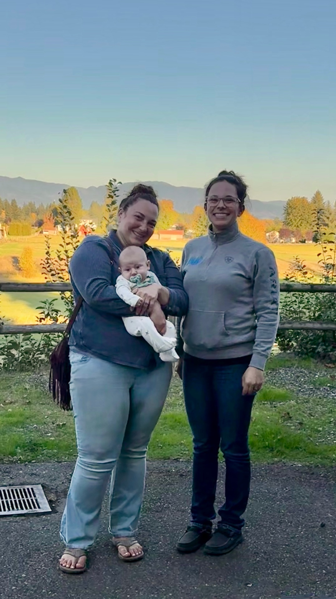 Two smiling guests standing together on the gravel by the pasture fence, one holding a ten-week-old baby, with golden autumn fields and mountains beyond