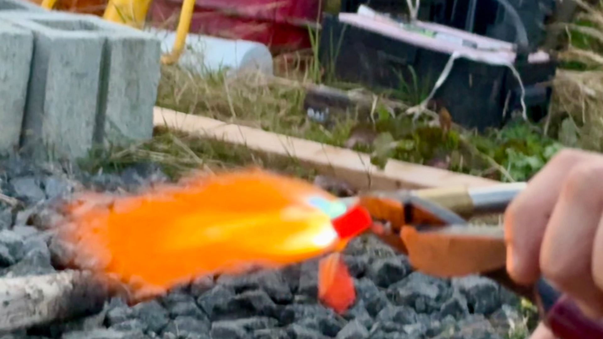 A glowing red-hot piece dropping away from the molten metal held in tongs, blazing orange in the torch flame