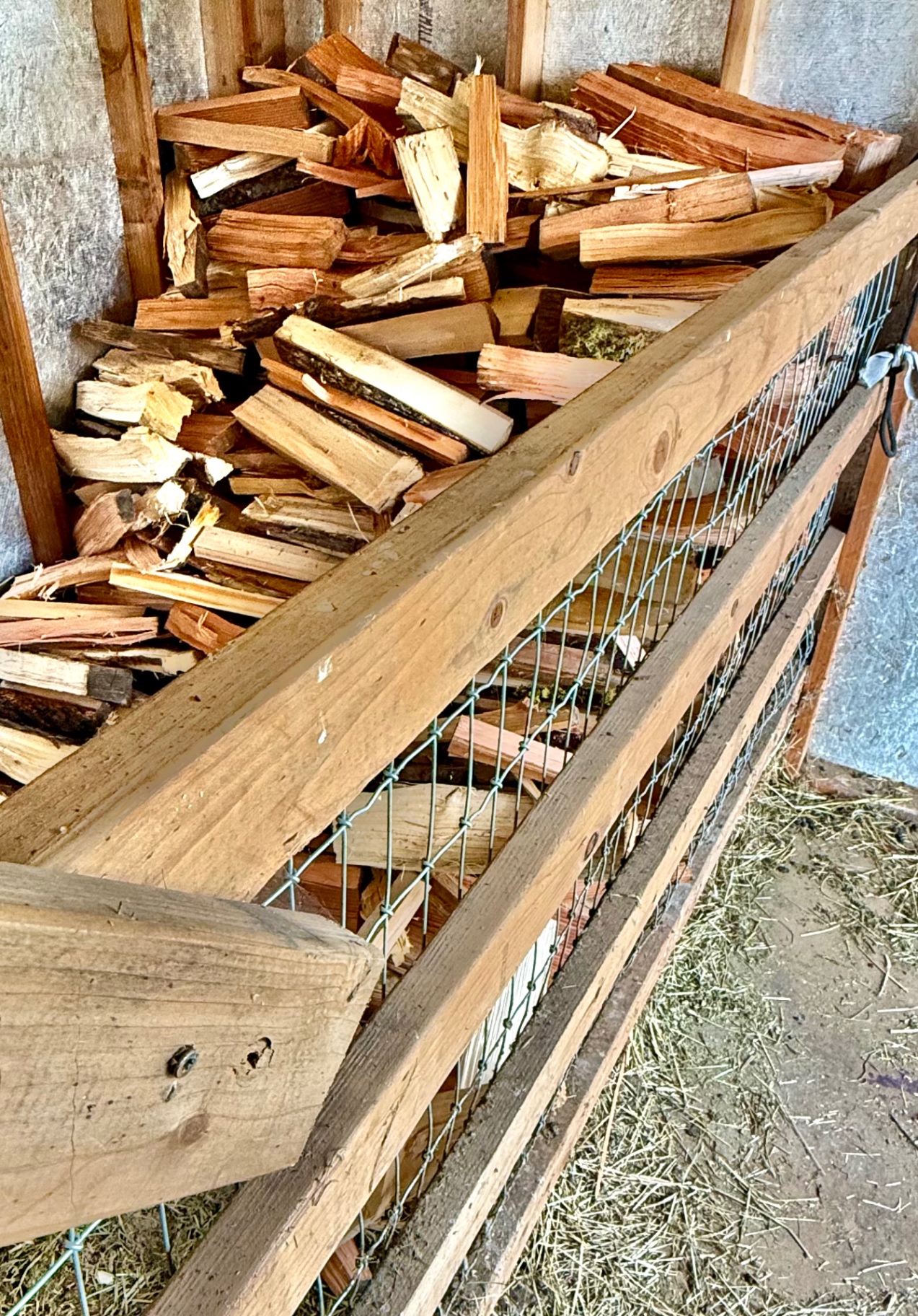 A close-up view into a wooden firewood bin filled with split firewood stacked behind a green wire-mesh railing inside a shed