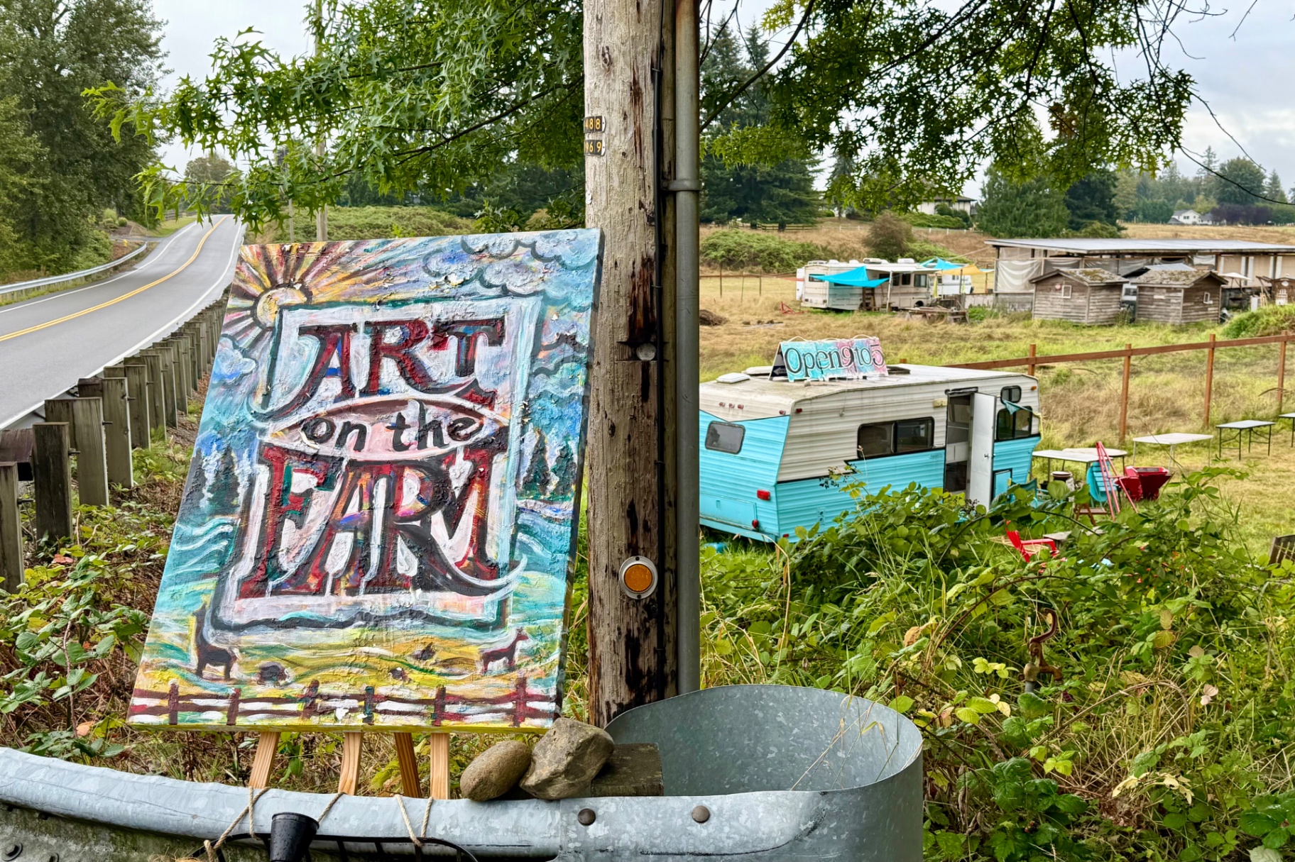 A hand-painted 'ART on the FARM' sign on an easel beside a road, with a turquoise trailer and outbuildings in a grassy field behind it