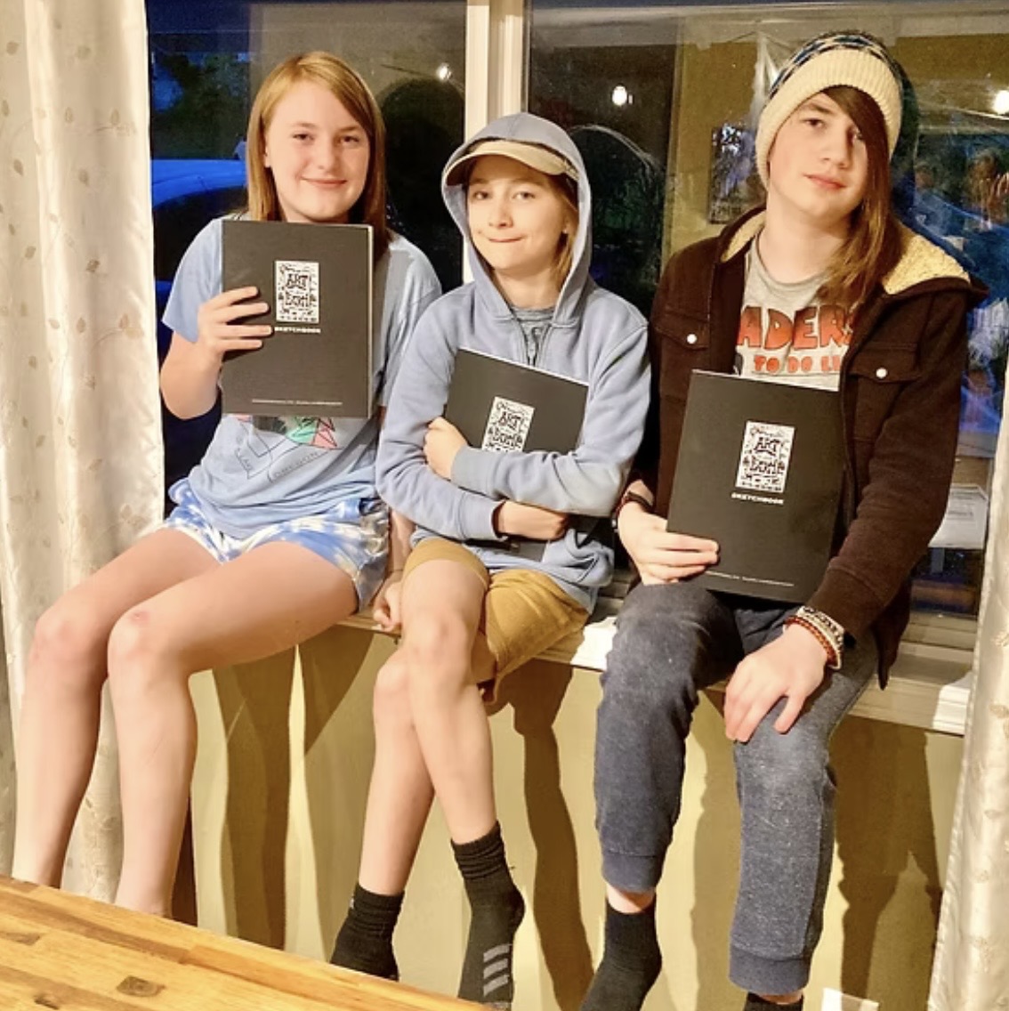 Three young artists each holding a black Art on the Farm sketchbook on a windowsill