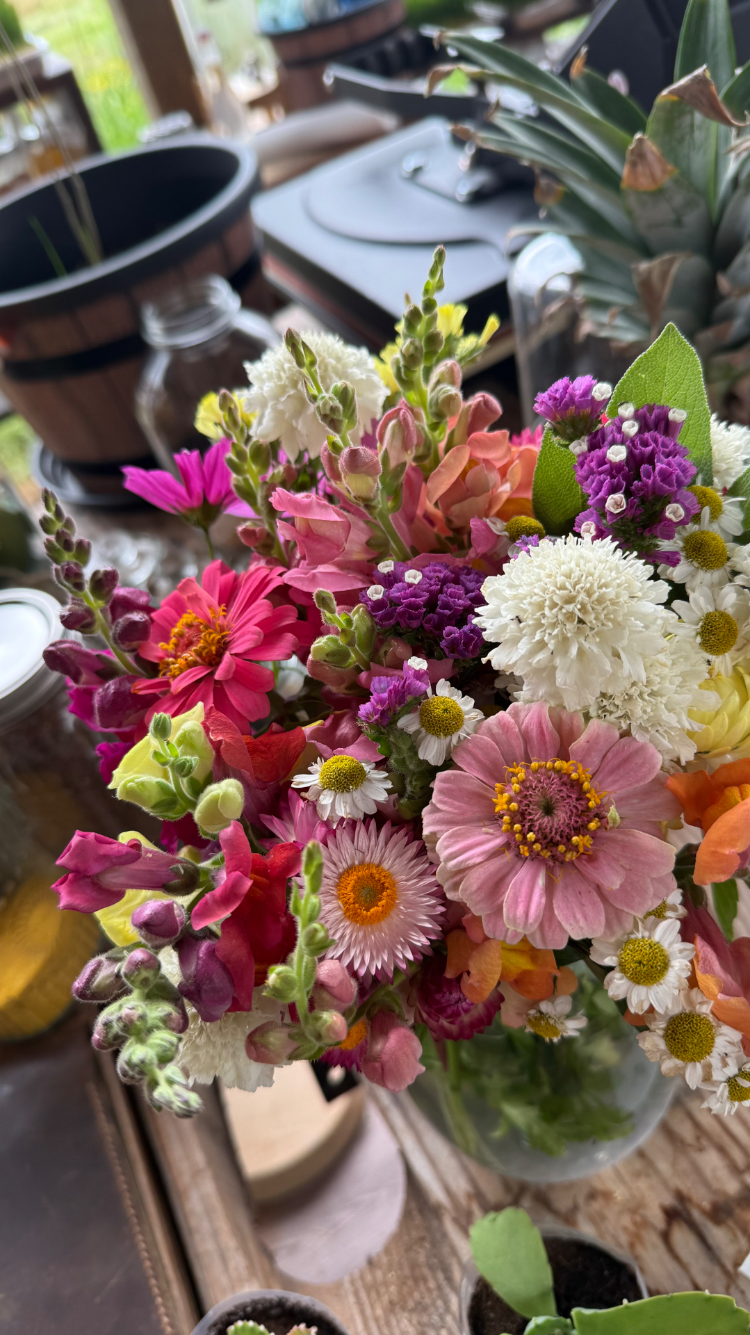 A lush multicolored garden bouquet of pink zinnias, magenta and orange snapdragons, purple statice, and a pale strawflower in a clear glass vase on a wooden surface