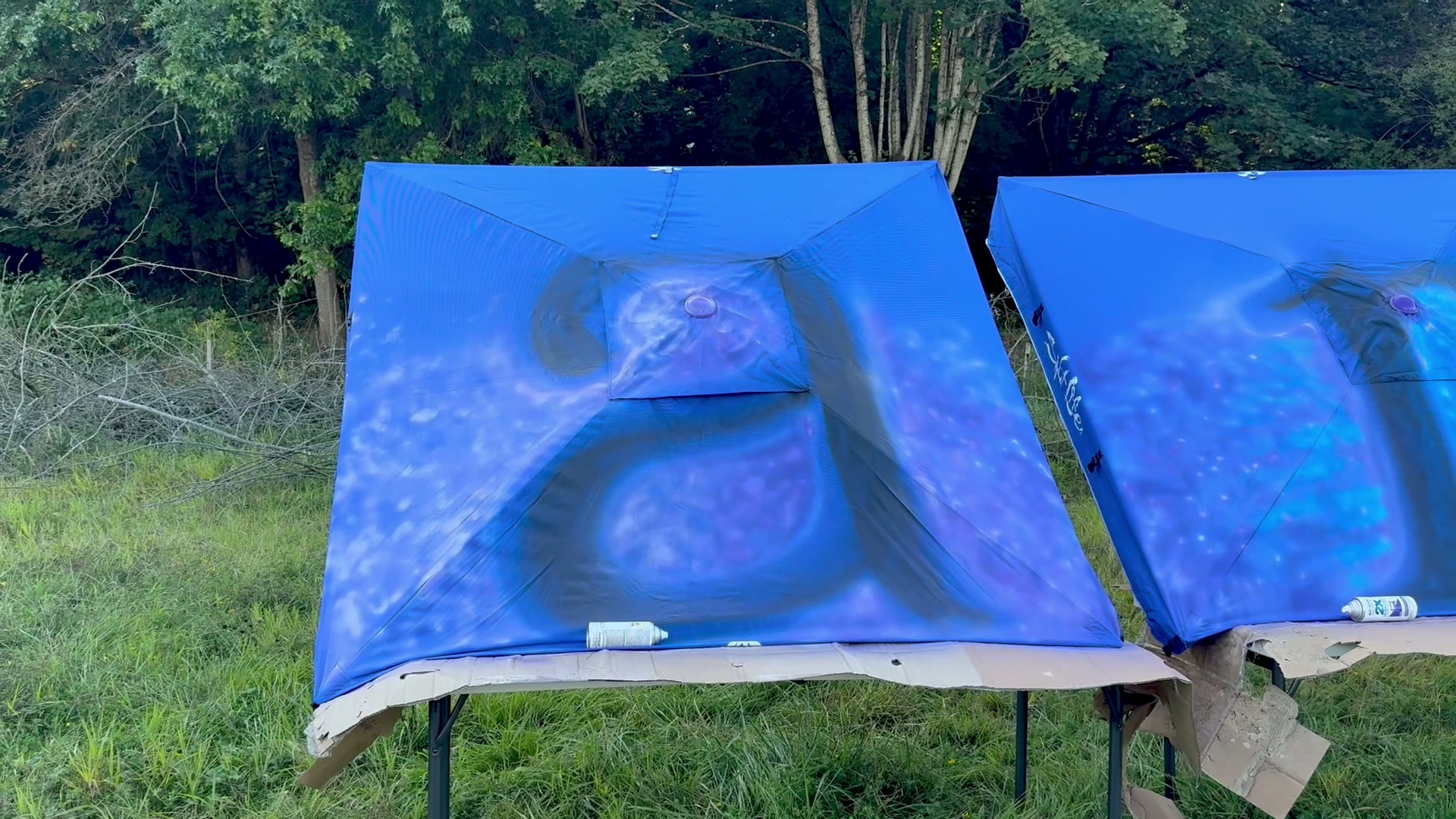 Two royal-blue canopy awnings spread out on a grassy field, being hand-painted with large stylized letters filled with purple and blue swirls of spray paint, spray cans resting on the edges