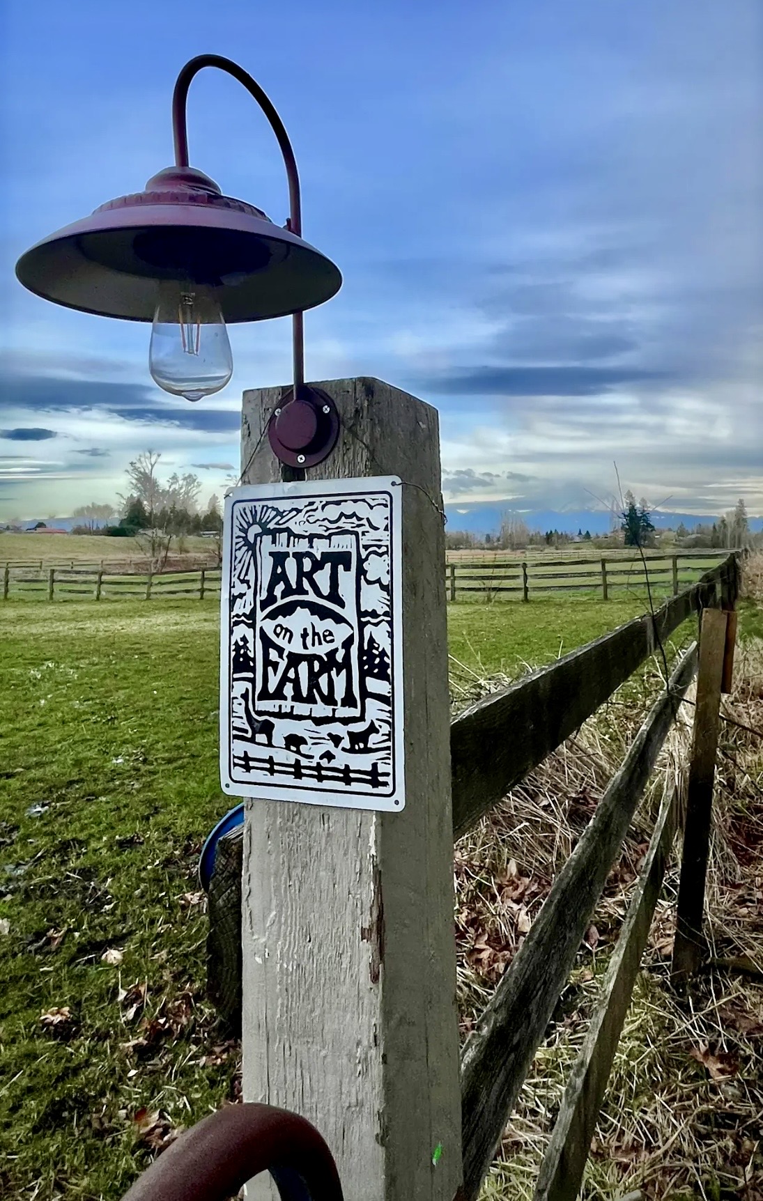 A weathered fence post at the farm entrance bearing an ART on the FARM sign, a maroon gooseneck lamp, and a cow-and-sheep graphic, with green pasture and mountains behind