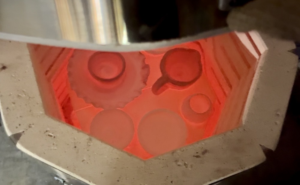 An overhead view into a glowing pottery kiln during firing, with bowls, cups, and plates silhouetted against the intense orange-red glow of the firebricks