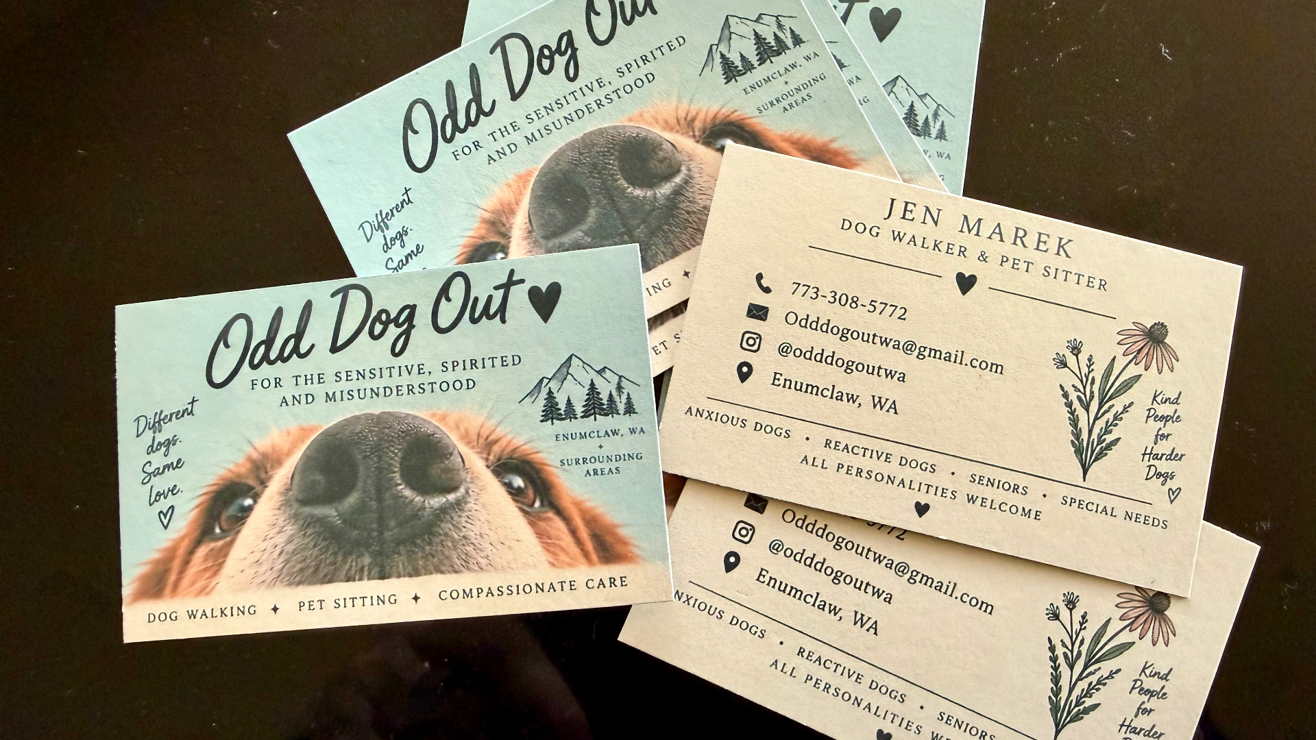 A set of finished double-sided business cards for 'Odd Dog Out' dog walking and pet sitting laid out on a dark surface, showing a sage-green front with a dog's face and a cream back with Jen Marek's contact details
