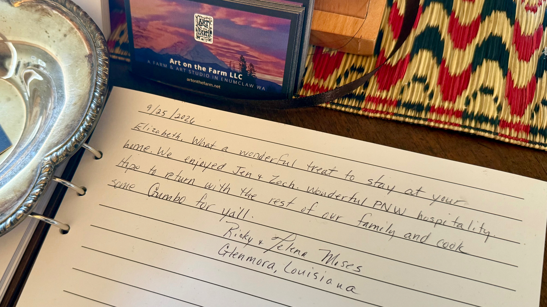 An open guest book with a handwritten note from Ricky and Jelena Moses, alongside business cards and a woven red mat on a dark wooden surface