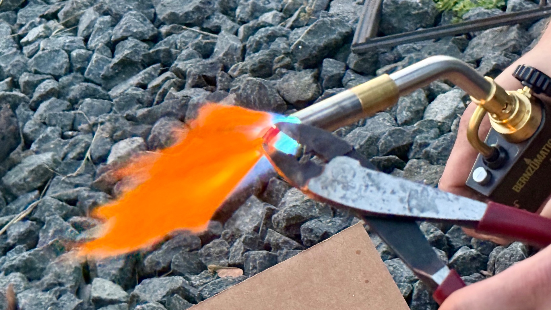 A close-up of a torch flame washing orange over a glowing piece of metal held in tongs over gravel