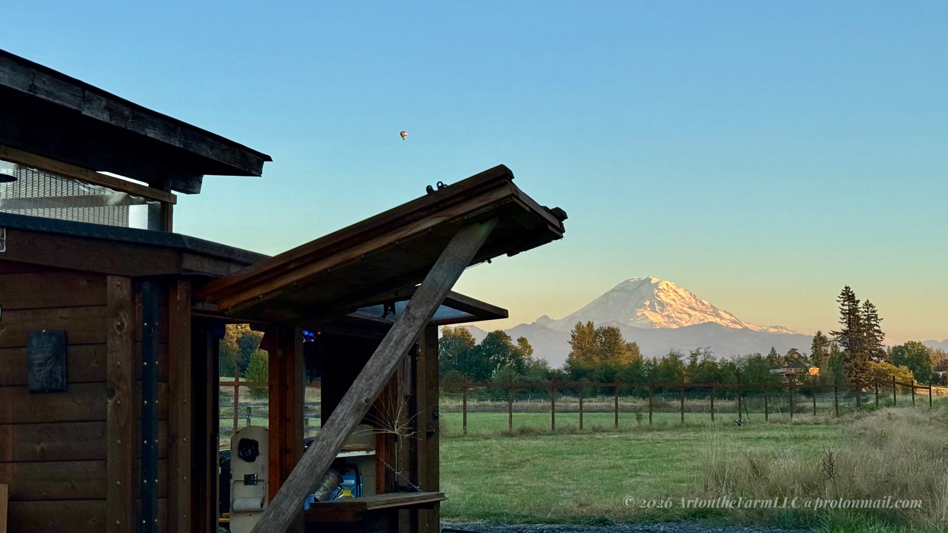 A snow-capped Mount Rainier glowing in soft light, framed by a dark wooden structure on the left, with a single small hot air balloon in the pale blue sky