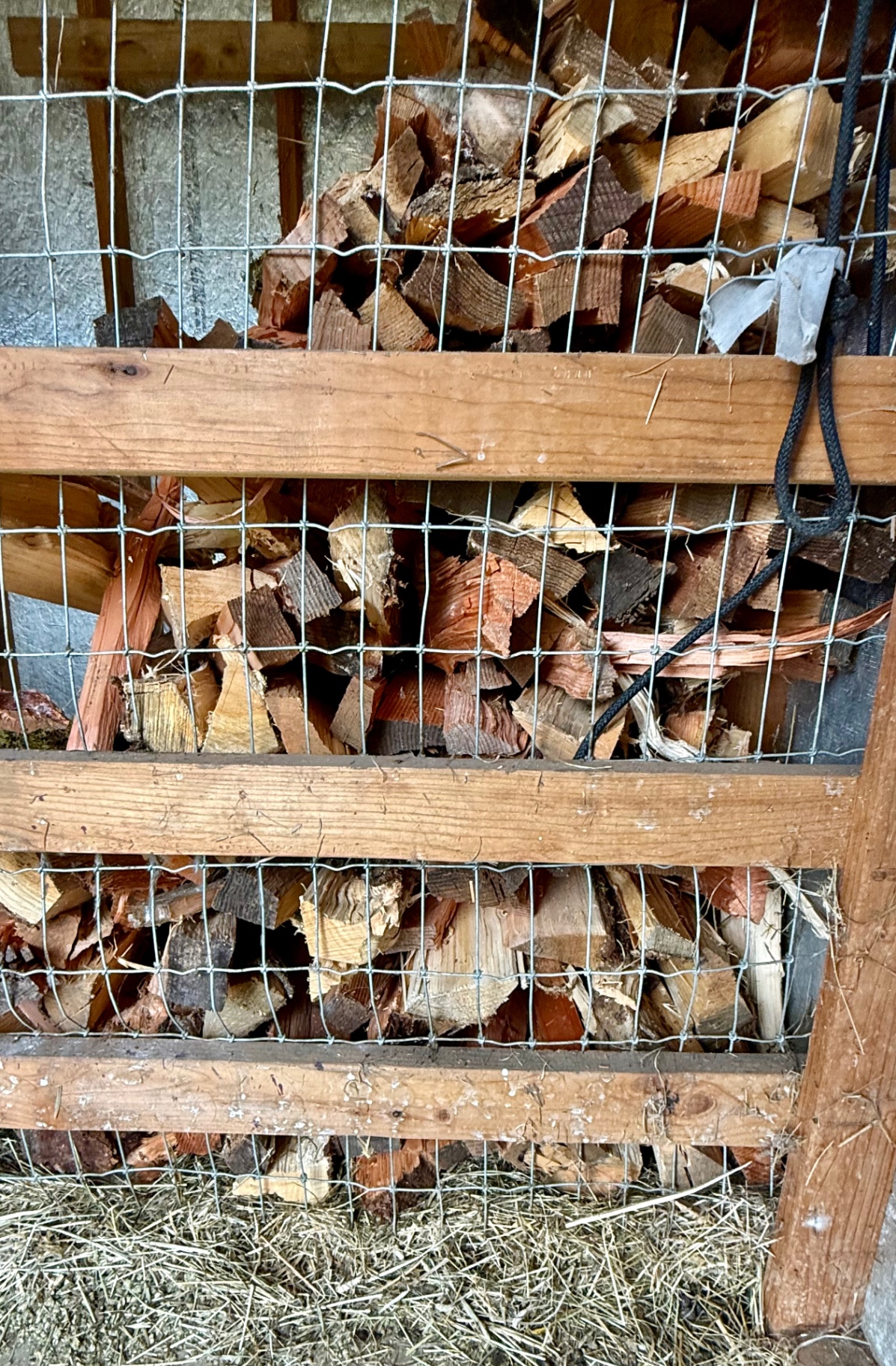 A vertical section of a firewood storage structure filled with split logs behind silver-grey wire mesh and wooden planks
