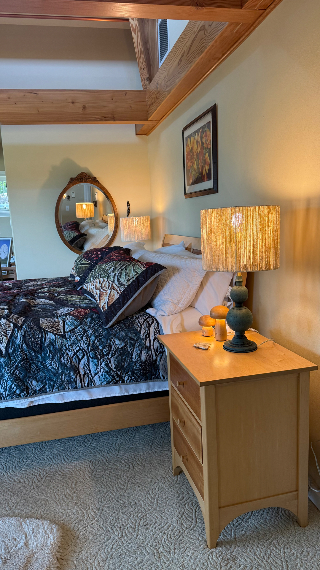 An angled close-up of a bedside nightstand with a dark-teal lamp and a glowing jute drum shade, two small glowing wooden mushroom lights beside it, and the tree-patterned quilt and a white pillow to the side