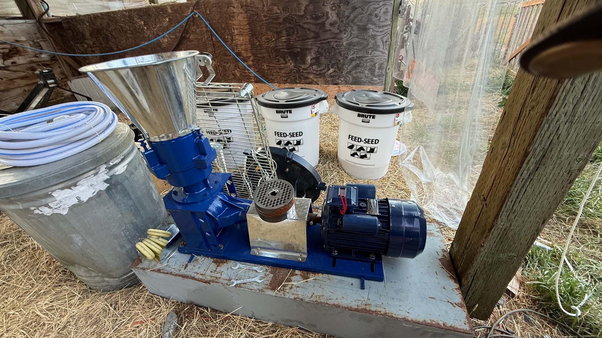 A blue pellet mill with hopper, die plate and motor beside FEED-SEED storage containers in a rustic work area