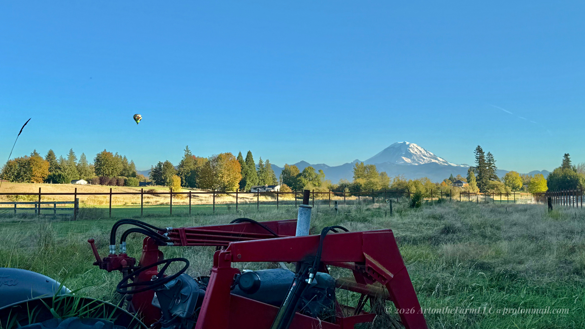 Mount Rainier rising beyond autumn trees and a fenced pasture, with a colorful hot air balloon in the blue sky and a red farm tractor in the foreground