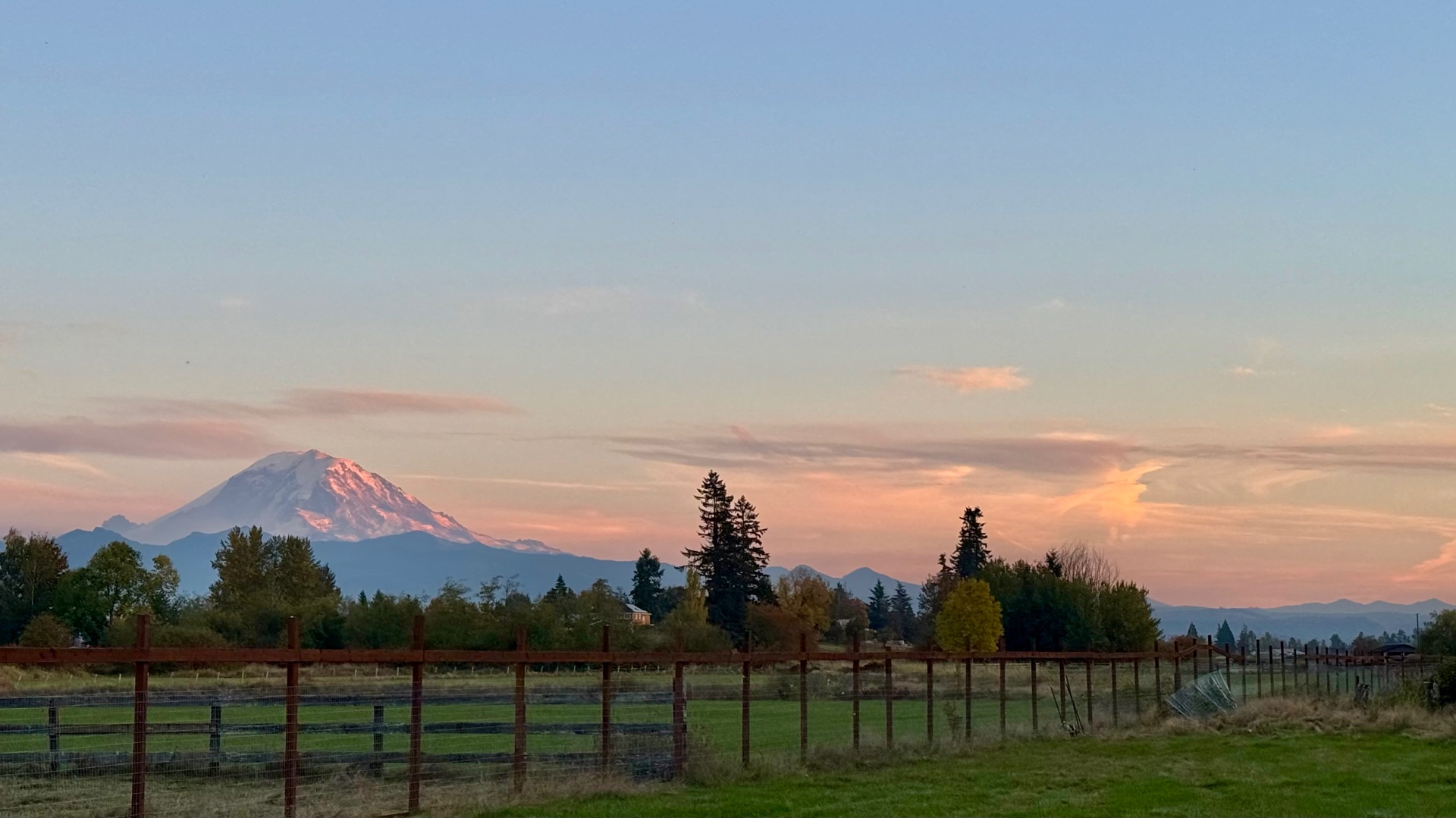Mount Rainier glowing pink in the late evening light above a green pasture lined with a wooden post-and-wire fence, with soft pink and peach sunset clouds