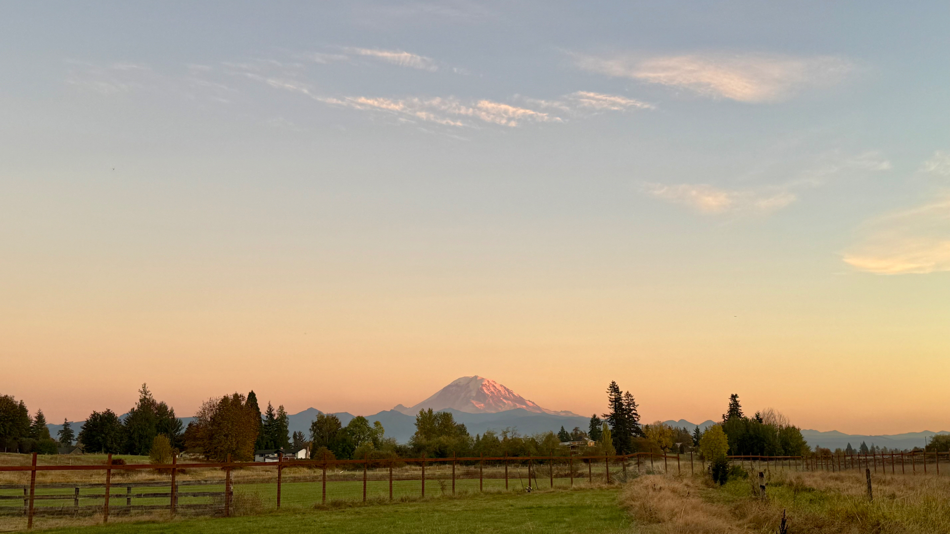 Mount Rainier at sunset above a line of autumn trees and a grassy pasture, a reddish-brown post-and-wire fence running across the field, the sky fading from pale blue to warm peach and rose