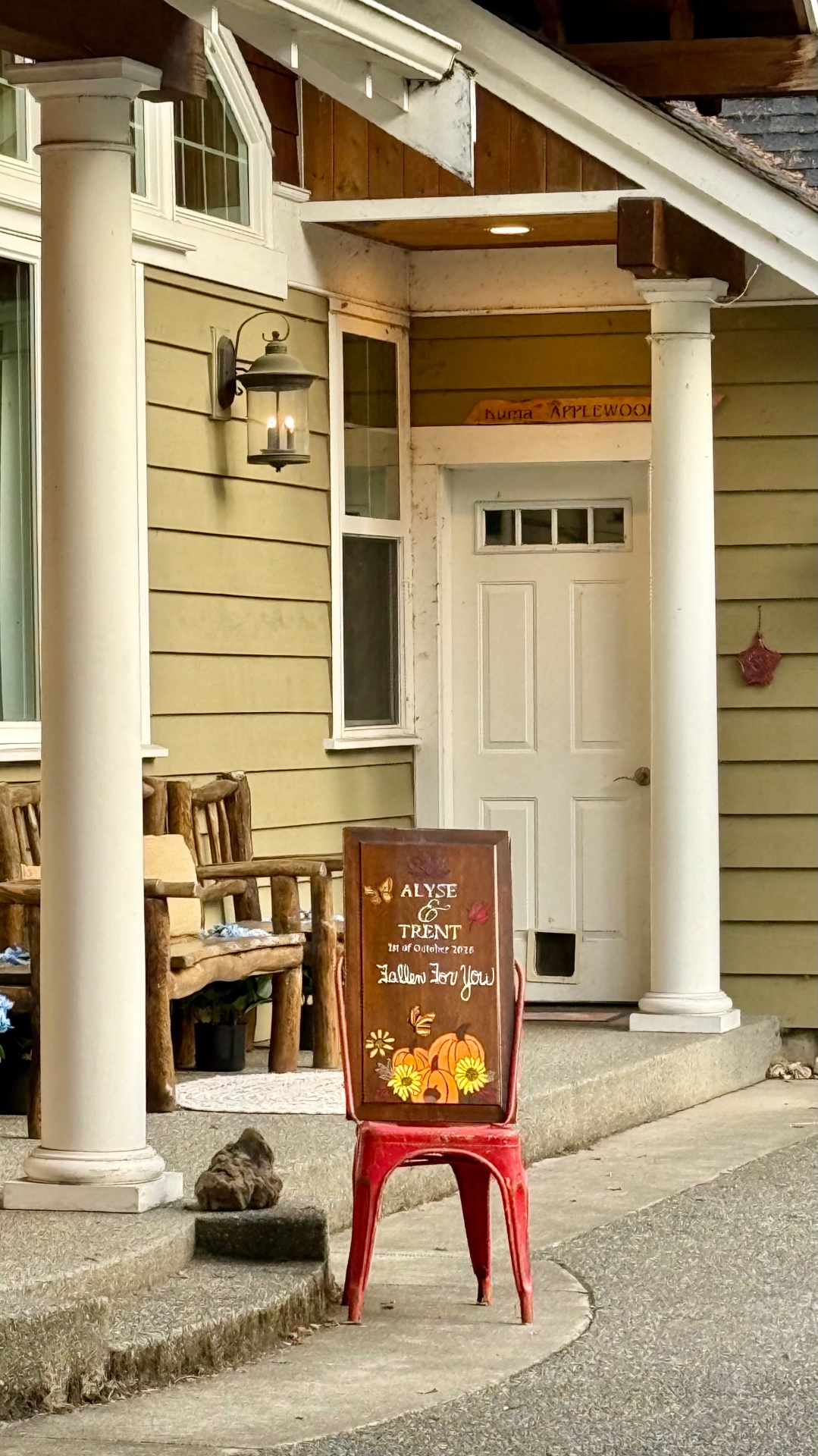 The wedding sign on a red metal chair by the farm's front porch, with white columns and a lit lantern above
