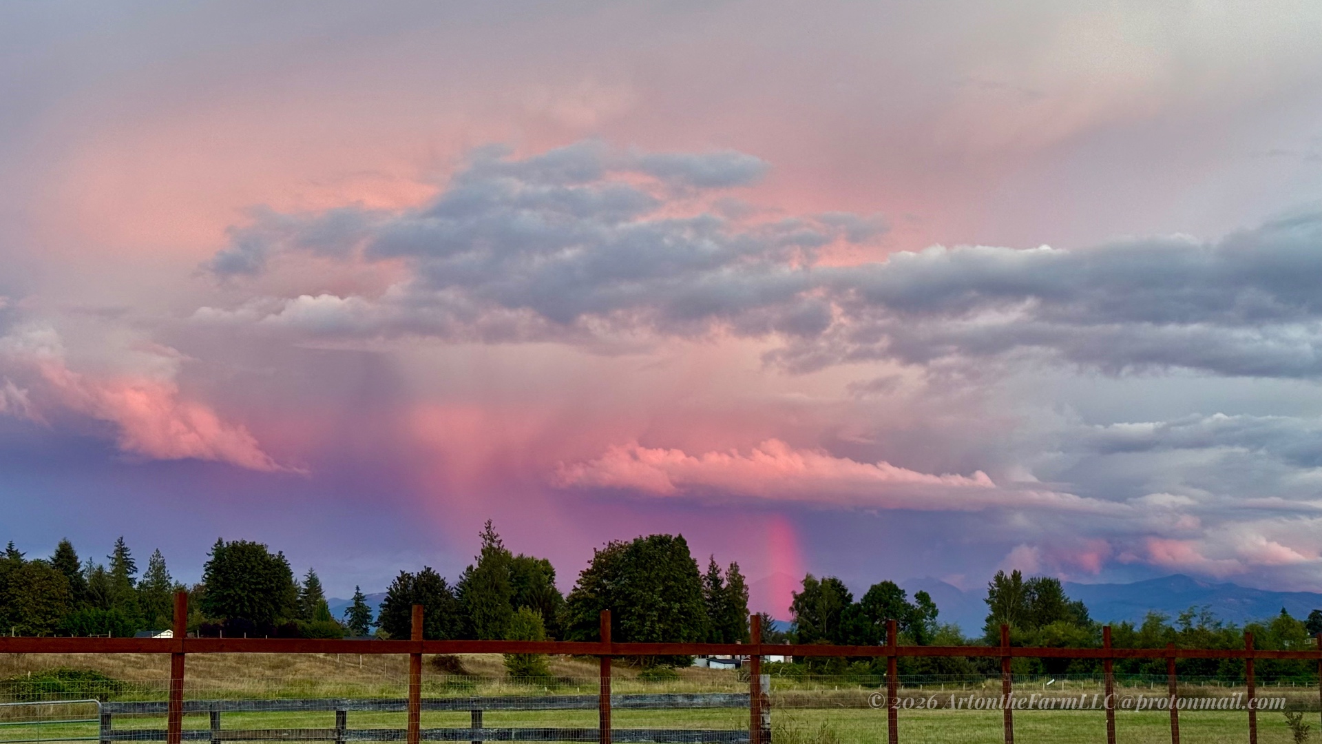 A dramatic magenta and pink sunset sky over a wooden fence, golden field, tree line, and distant blue-grey mountains at twilight
