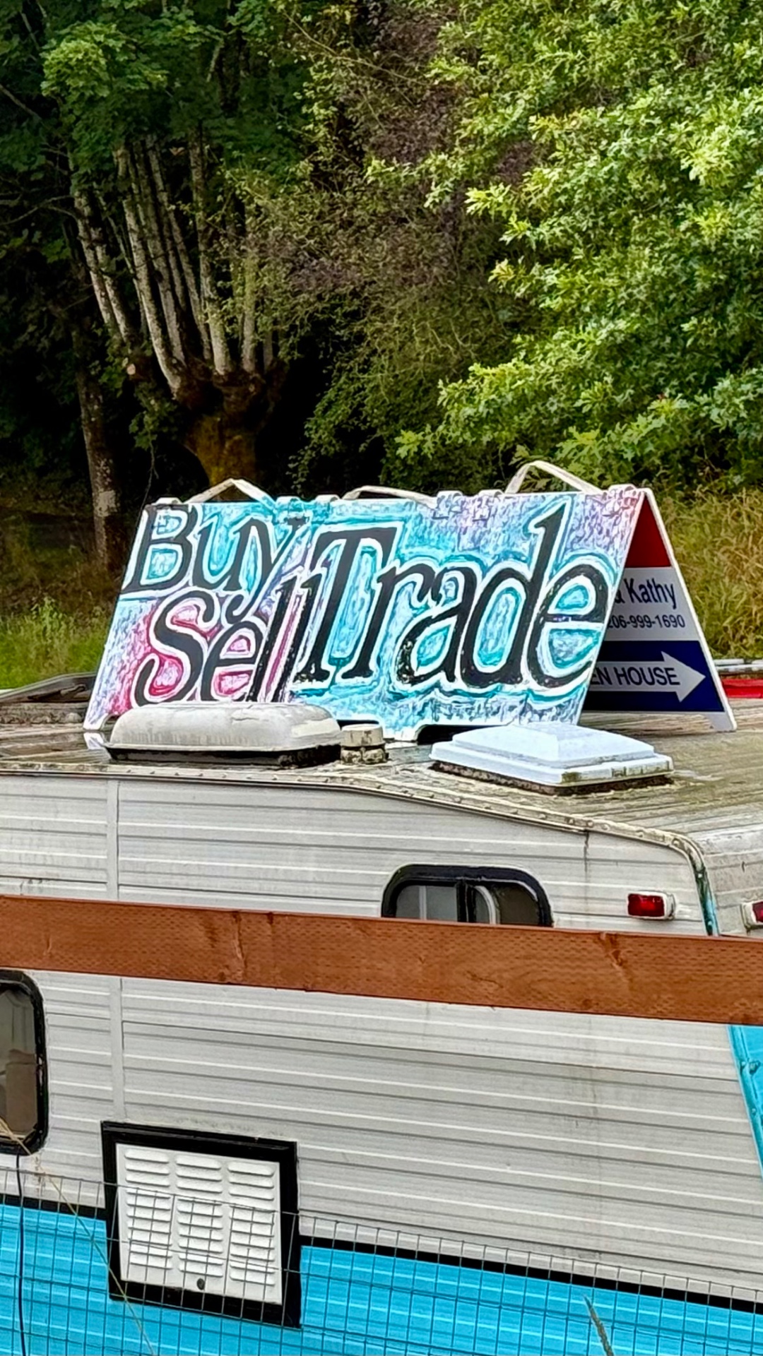 Close-up of a colorful hand-painted 'Buy Sell Trade' sign mounted on the roof of a turquoise and white vintage trailer