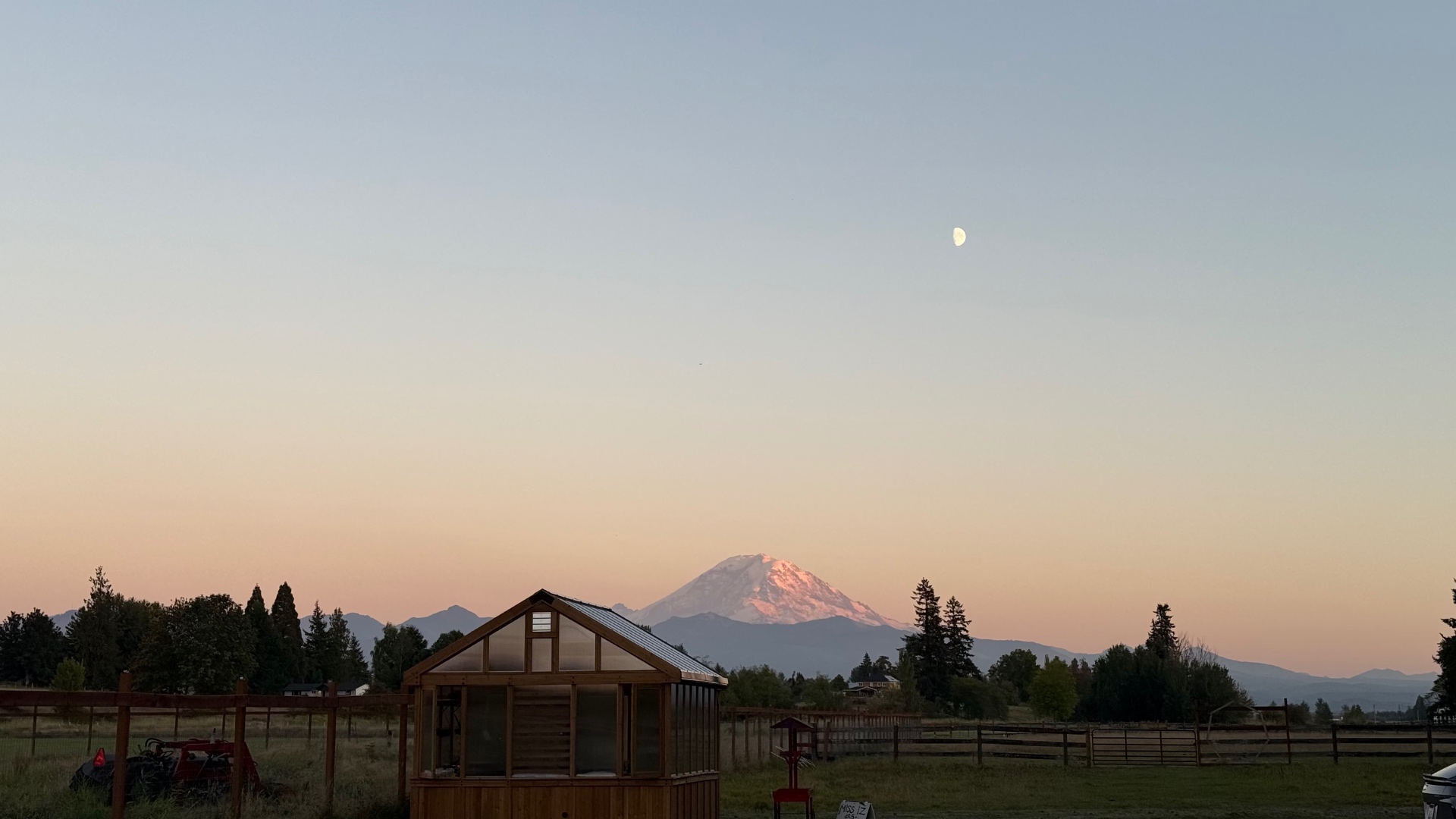 A wide twilight view of snow-capped Mount Rainier glowing pink in the last sunset light, with a half moon in a soft peach-and-blue sky and a small wooden greenhouse in the foreground