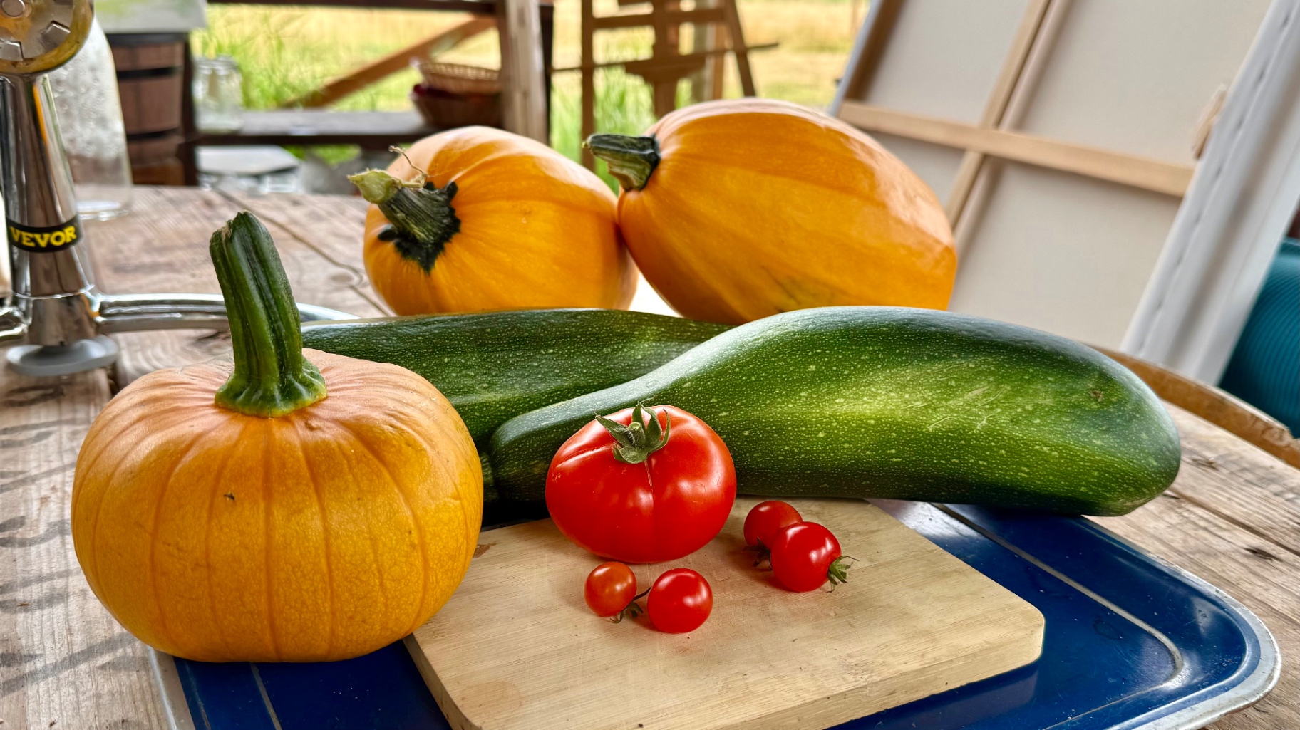 Orange pumpkins, dark green zucchini, and red tomatoes on a cutting board atop a blue tray with a metal juicer press on a wooden table