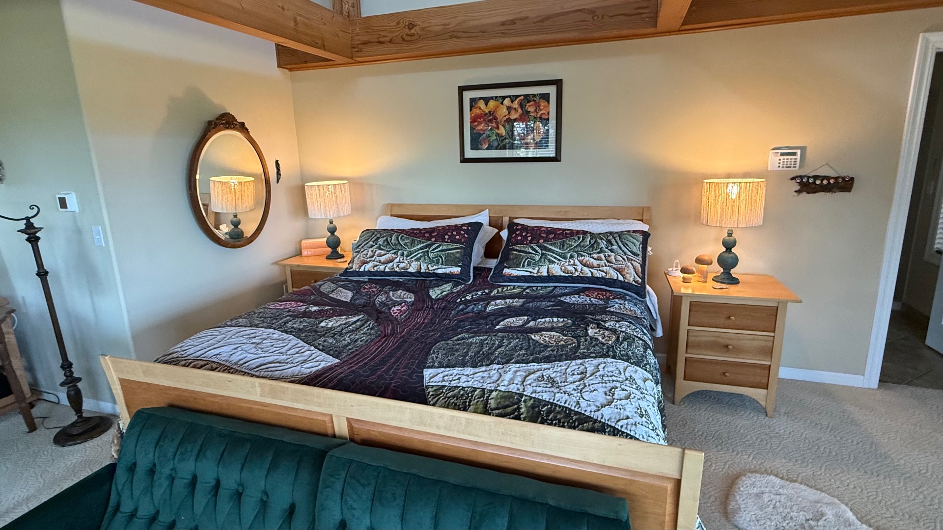 A wide evening view of a master bedroom with a light-wood bed under a tree-motif quilt, two matching nightstands with turned-on jute-shaded lamps glowing warm yellow, exposed wood ceiling beams and a framed flower print above the bed