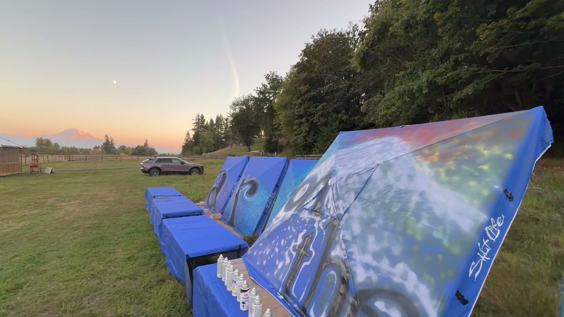 Blue painted canopy awnings set up across a field at dusk, rows of spray cans on a covered table in front, a crescent moon and pink sunset light on the mountain behind them