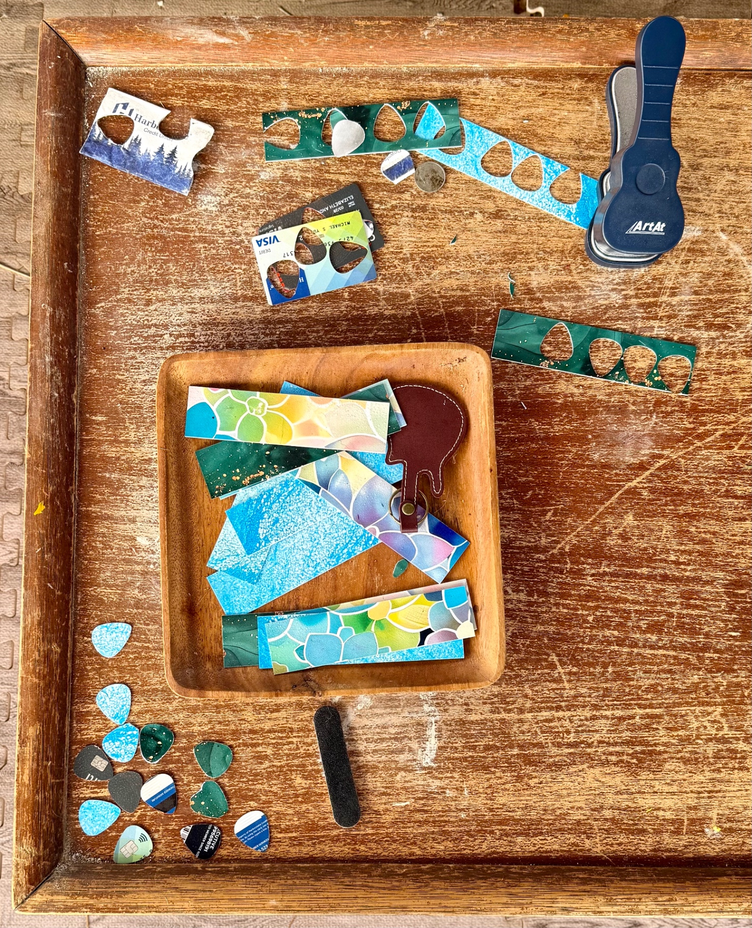 A wooden craft tray holding plastic card strips, a blue pick punch tool and finished guitar picks, with a sanding block and leather pick-holder keychain