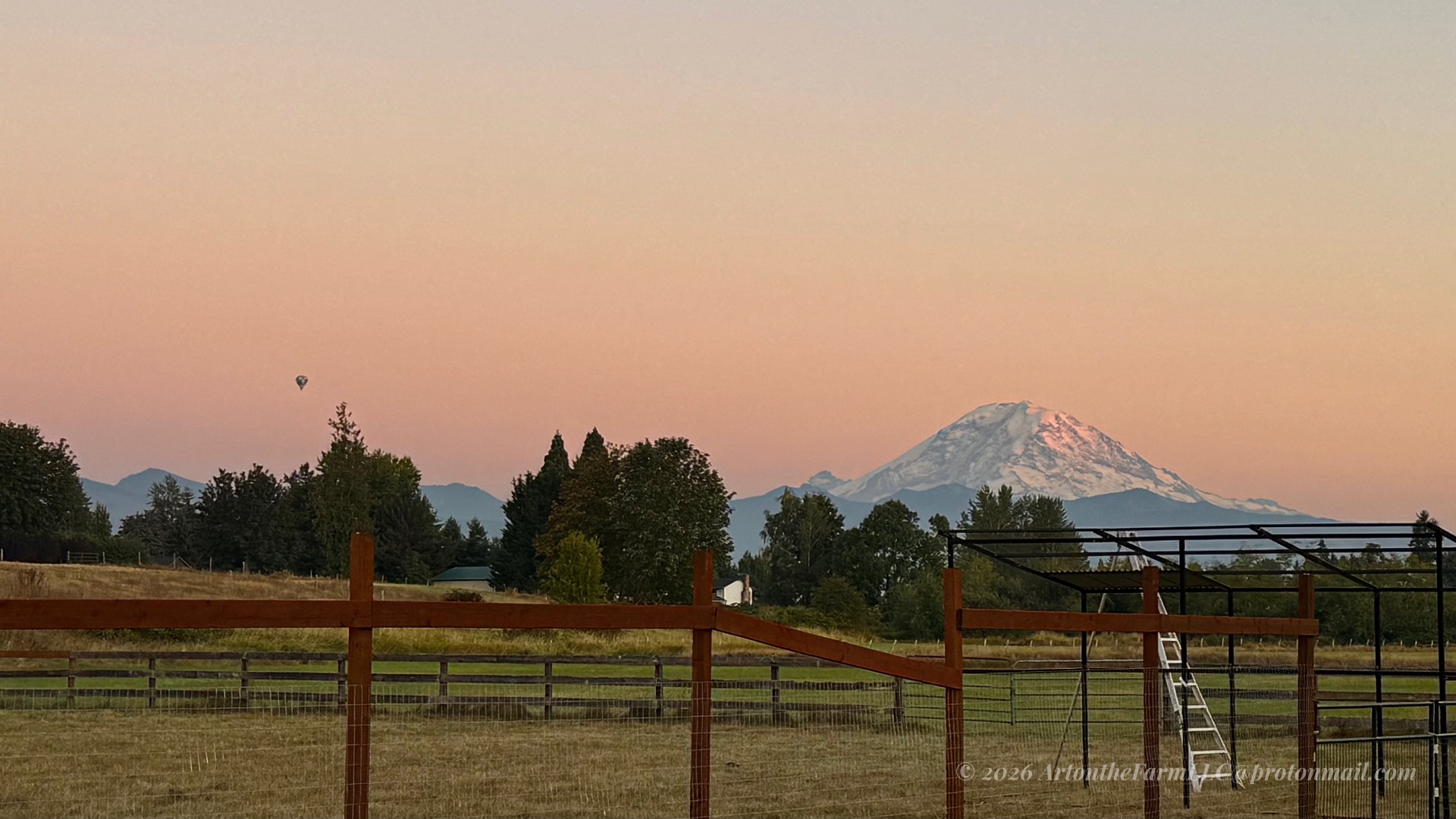 A pink-and-orange dawn sky over snow-capped Mount Rainier, with a wooden fence in the foreground and a single hot air balloon silhouette against the warm sky