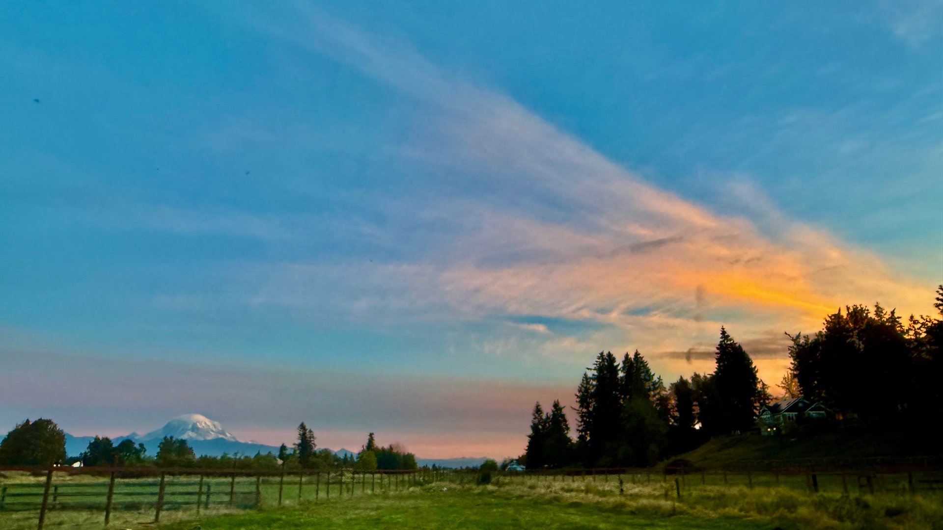 Wispy orange and pink sunset clouds over the farm pasture and evergreen trees, with Mount Rainier small in the distance and the farmhouse lights coming on