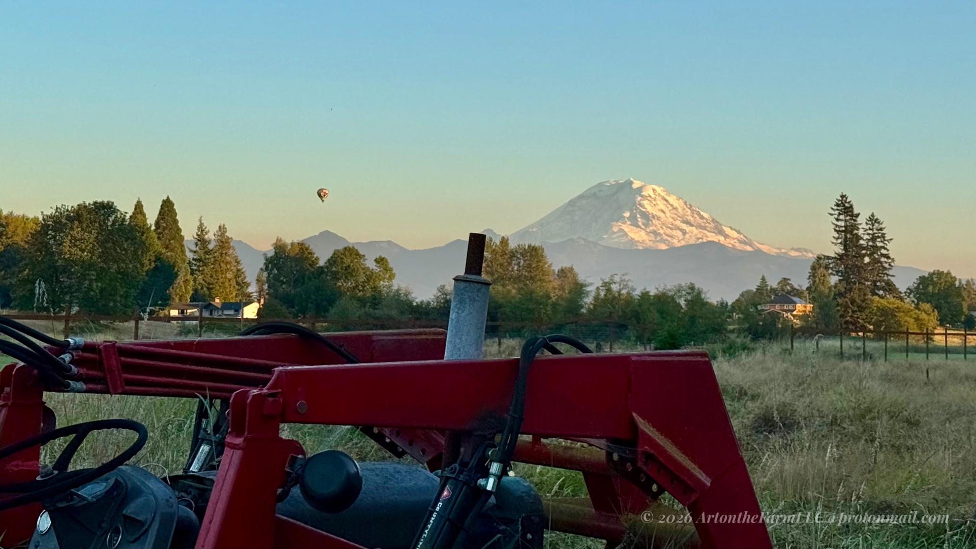 The red metal frame and black hydraulic hoses of a farm tractor in the foreground, with snow-capped Mount Rainier and a small hot air balloon in the distance