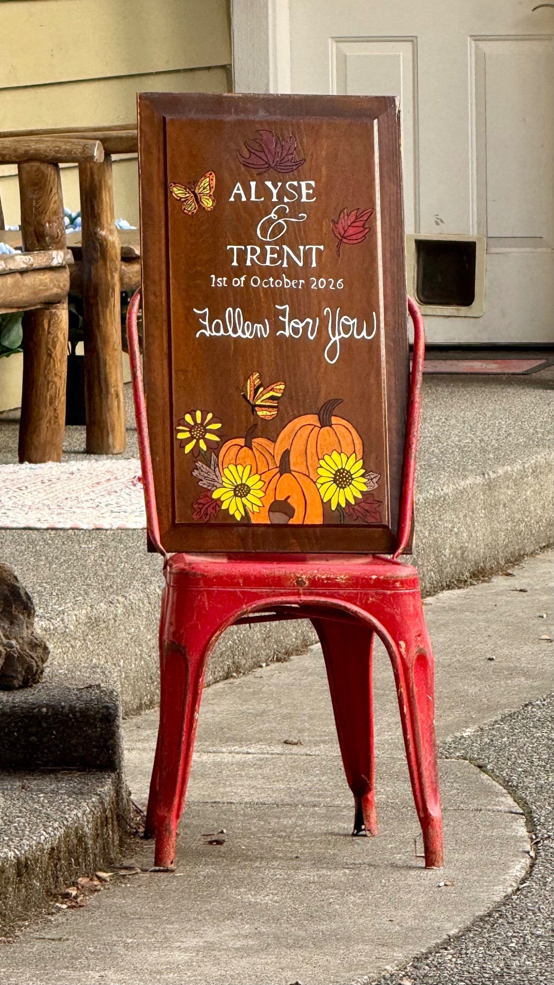 A close view of the hand-painted wooden sign reading “Alyse & Trent, 1st of October 2026 — Fallen For You,” decorated with pumpkins, sunflowers and butterflies