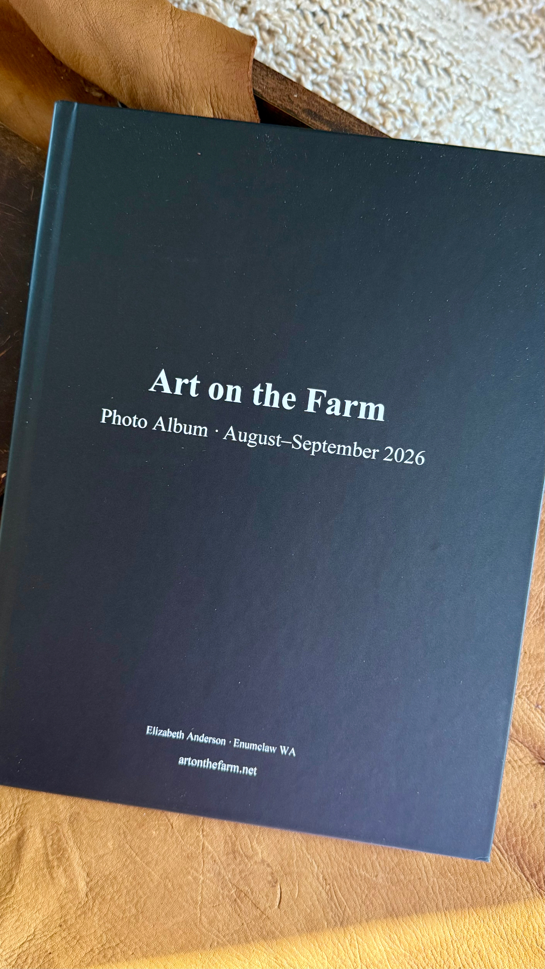 A black hardcover photo album reading Art on the Farm Photo Album August–September 2026, printed with Elizabeth Anderson Enumclaw WA and artonthefarm.net, lying on a leather cushion