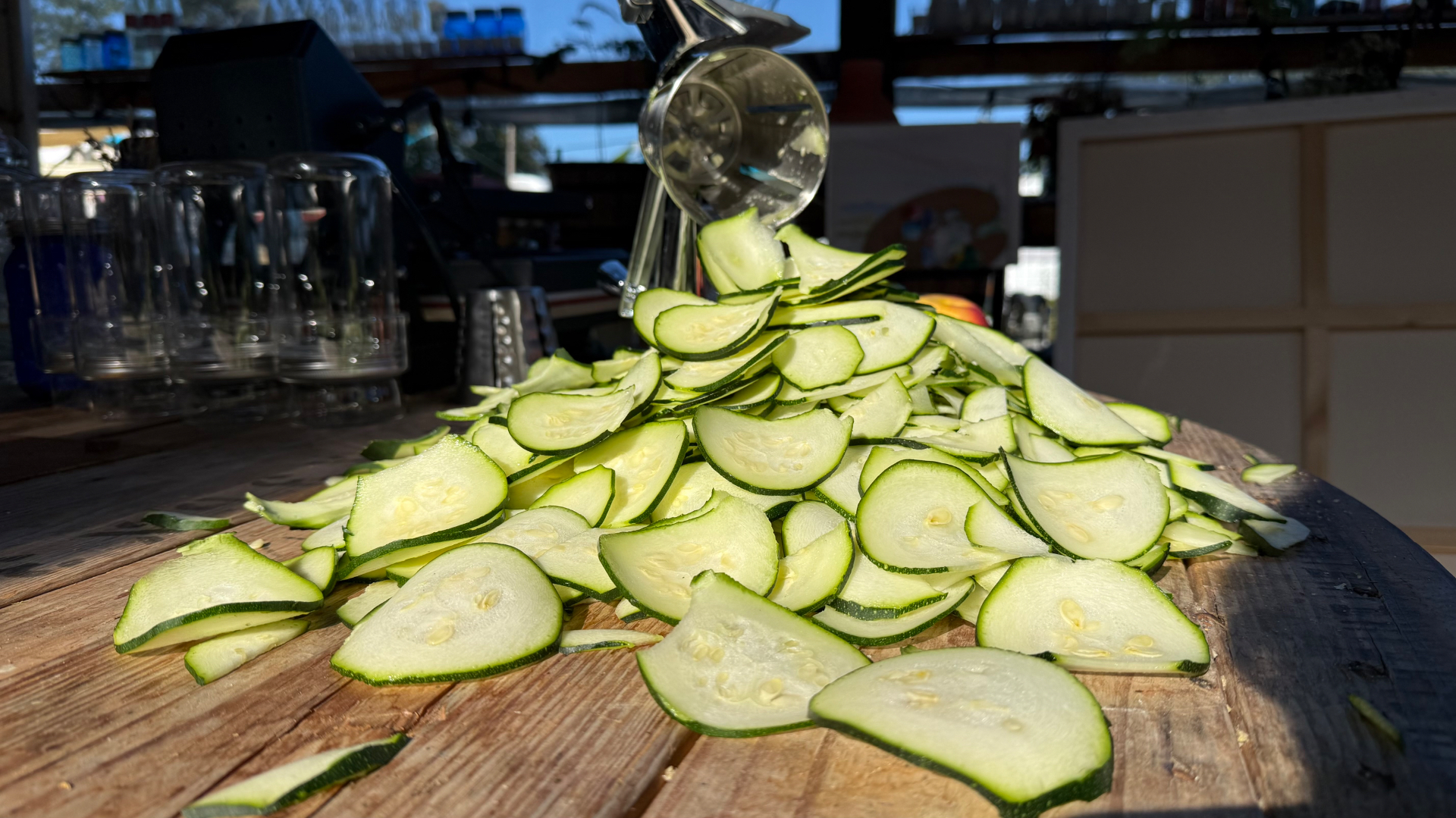 A large pile of thinly sliced pale zucchini rounds on a rustic wooden table with a silver rotary slicer and glass jars behind