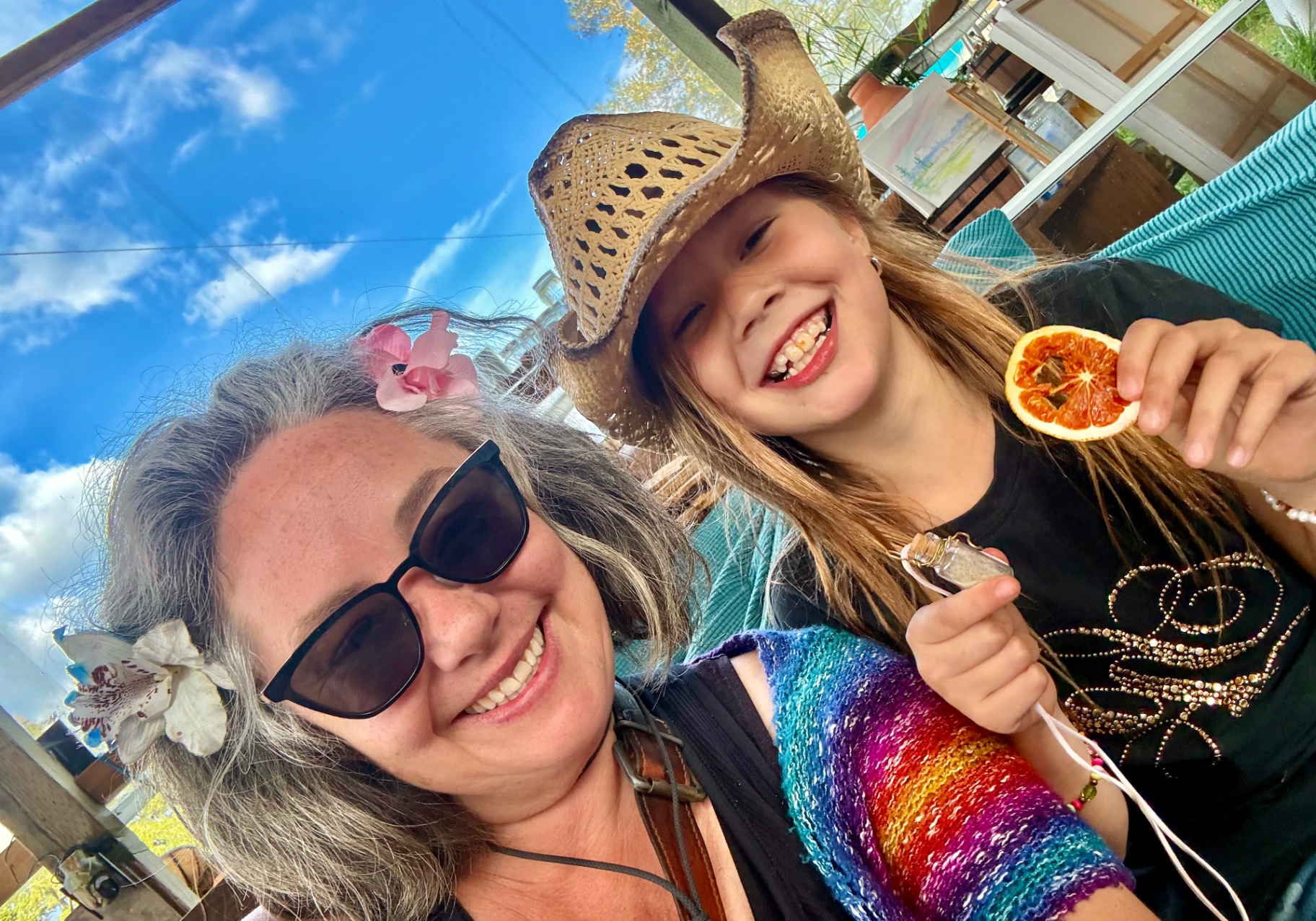 A smiling artist and a young camper in a straw hat holding dried fruit and a vial, on the sunlit farm studio porch