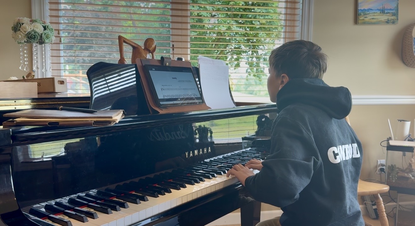 A young student seated at a glossy black grand piano, reading sheet music from a tablet in a sunlit room with trees visible through the window