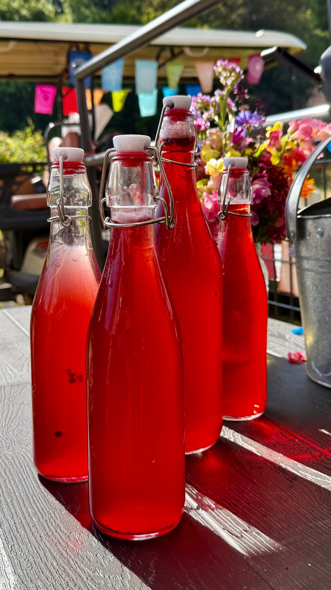 Four clear glass swing-top bottles filled with vibrant red liquid with white ceramic stoppers on a dark wooden table outdoors