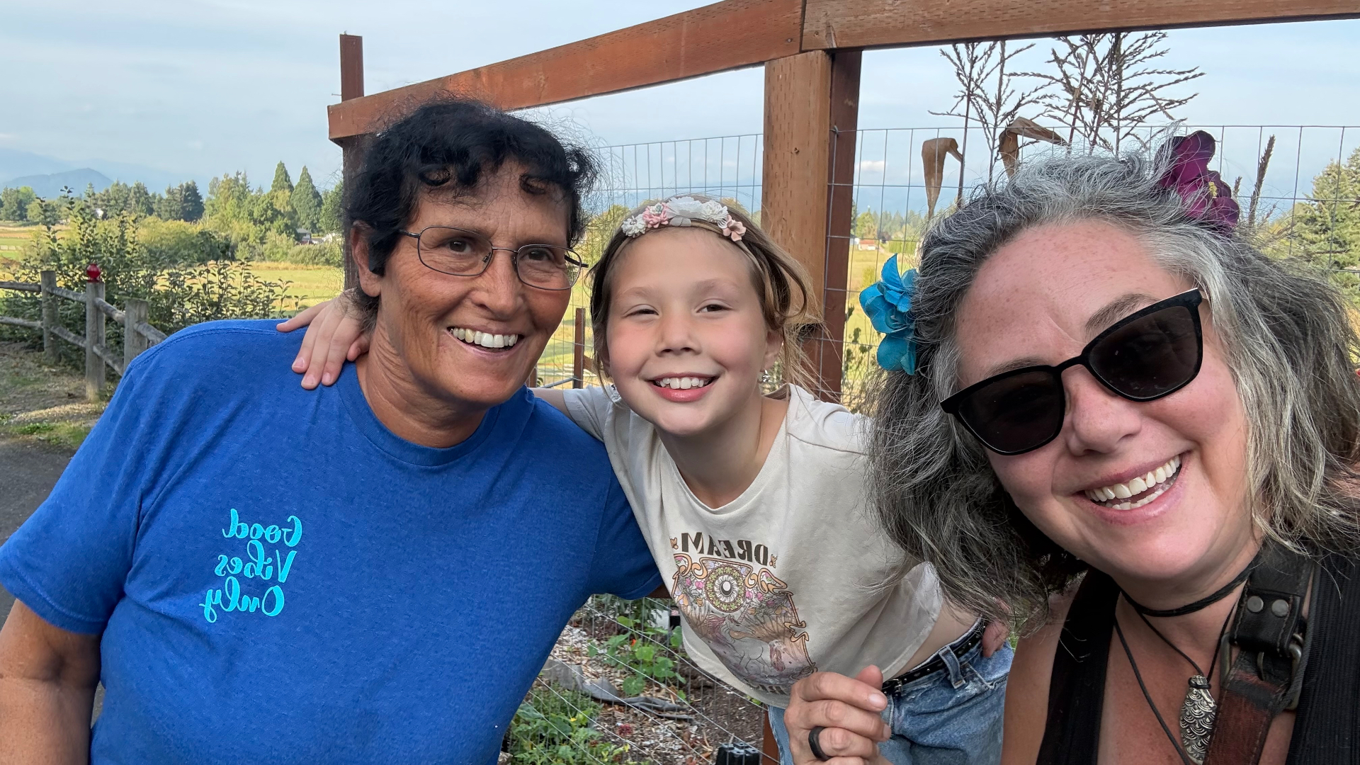 A smiling selfie on the farm — Miss Iz, Nanna and her granddaughter — posed against a wooden fence with green fields and hazy mountains behind