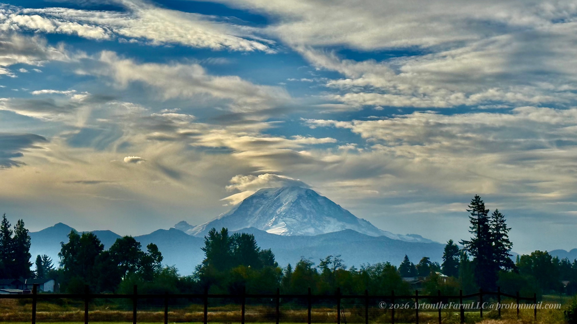 Mount Rainier beneath sweeping layers of autumn clouds, with the farm fence and trees in the foreground